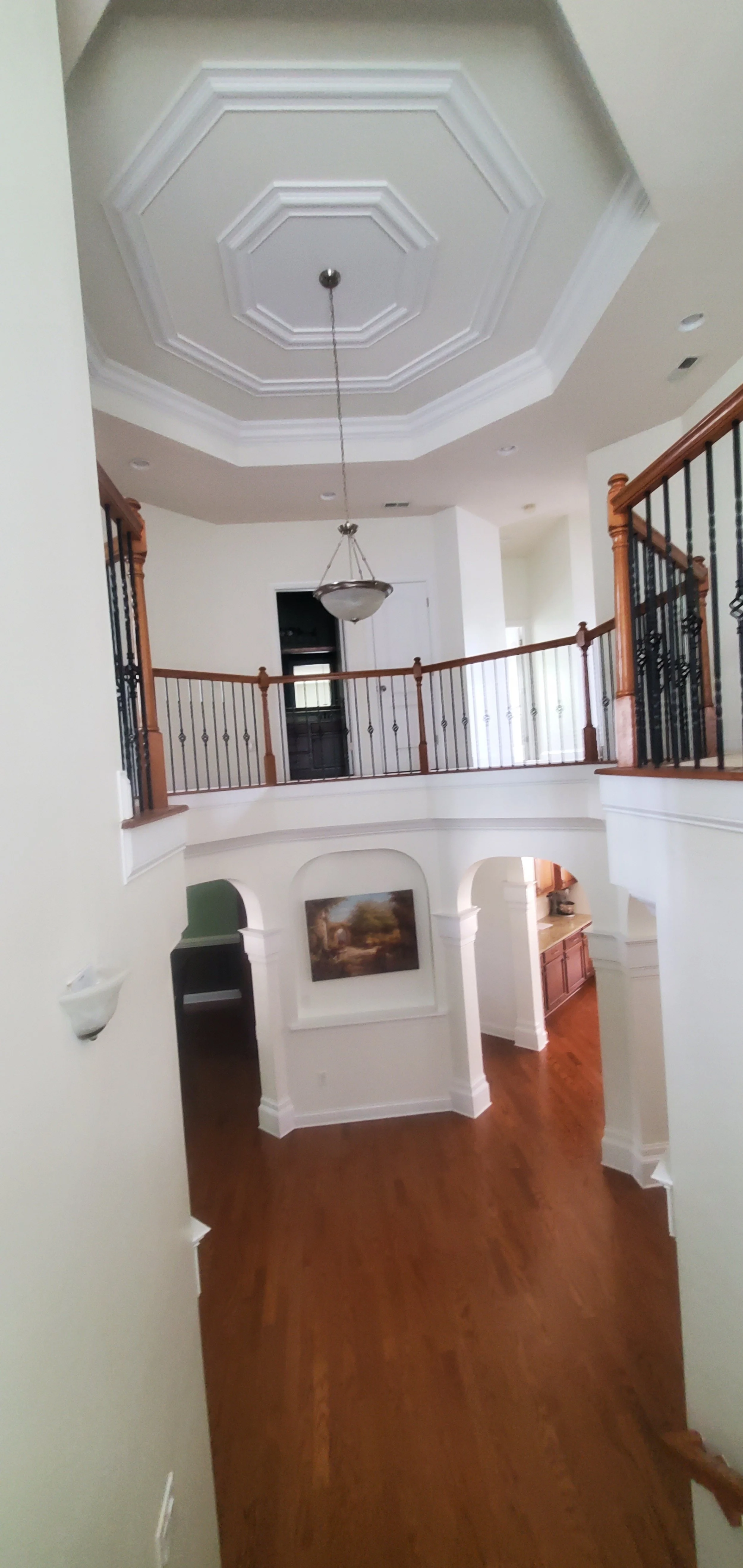 Interior view of a spacious, multi-level home with hardwood floors, white walls, and decorative ceiling molding. There is a hanging light fixture and a balcony with wooden and metal railing overlooking the lower floor. A framed painting is on the wal