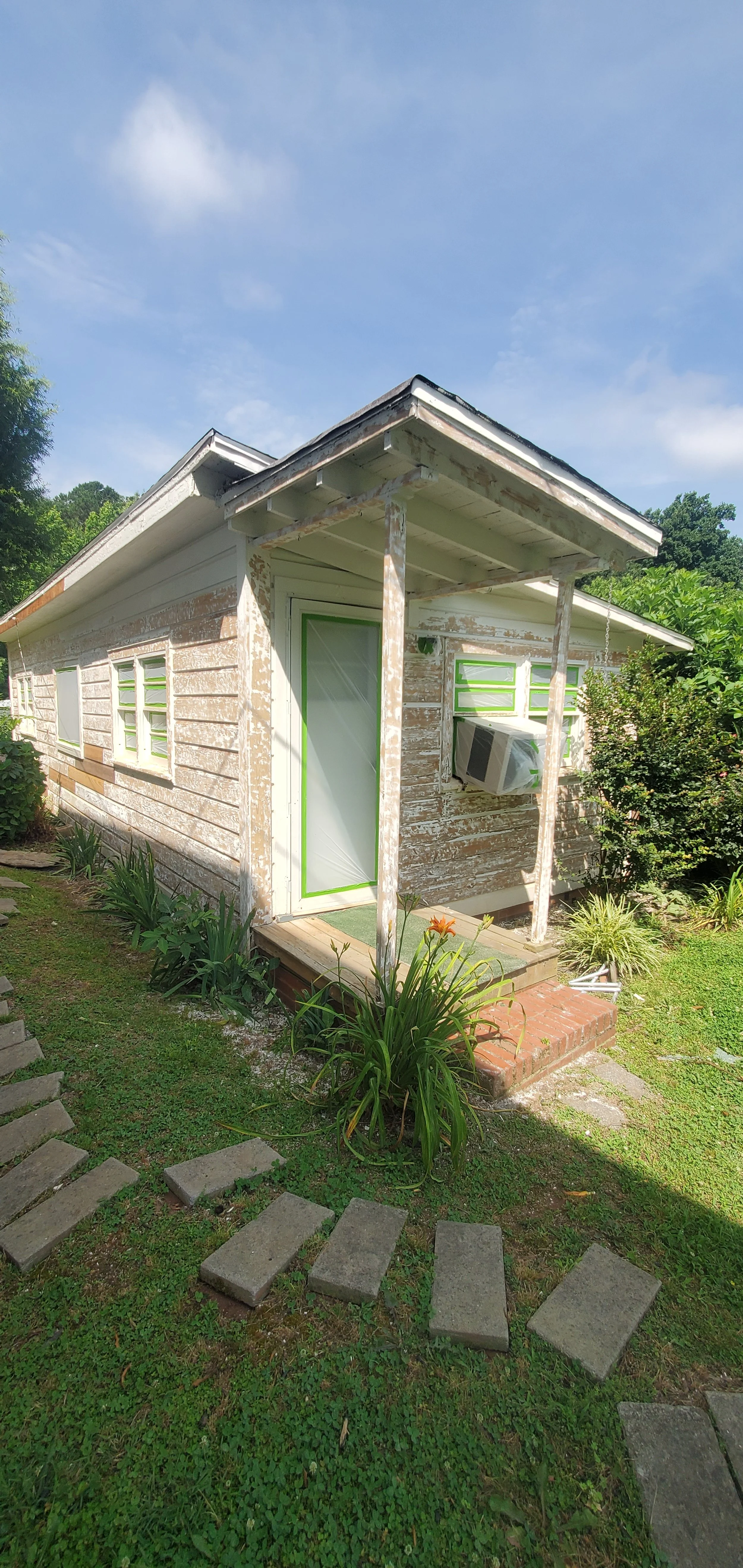 A small, weathered, wooden house with peeling paint, green trim, and an air conditioning unit in the window, surrounded by lush greenery and a clear blue sky.