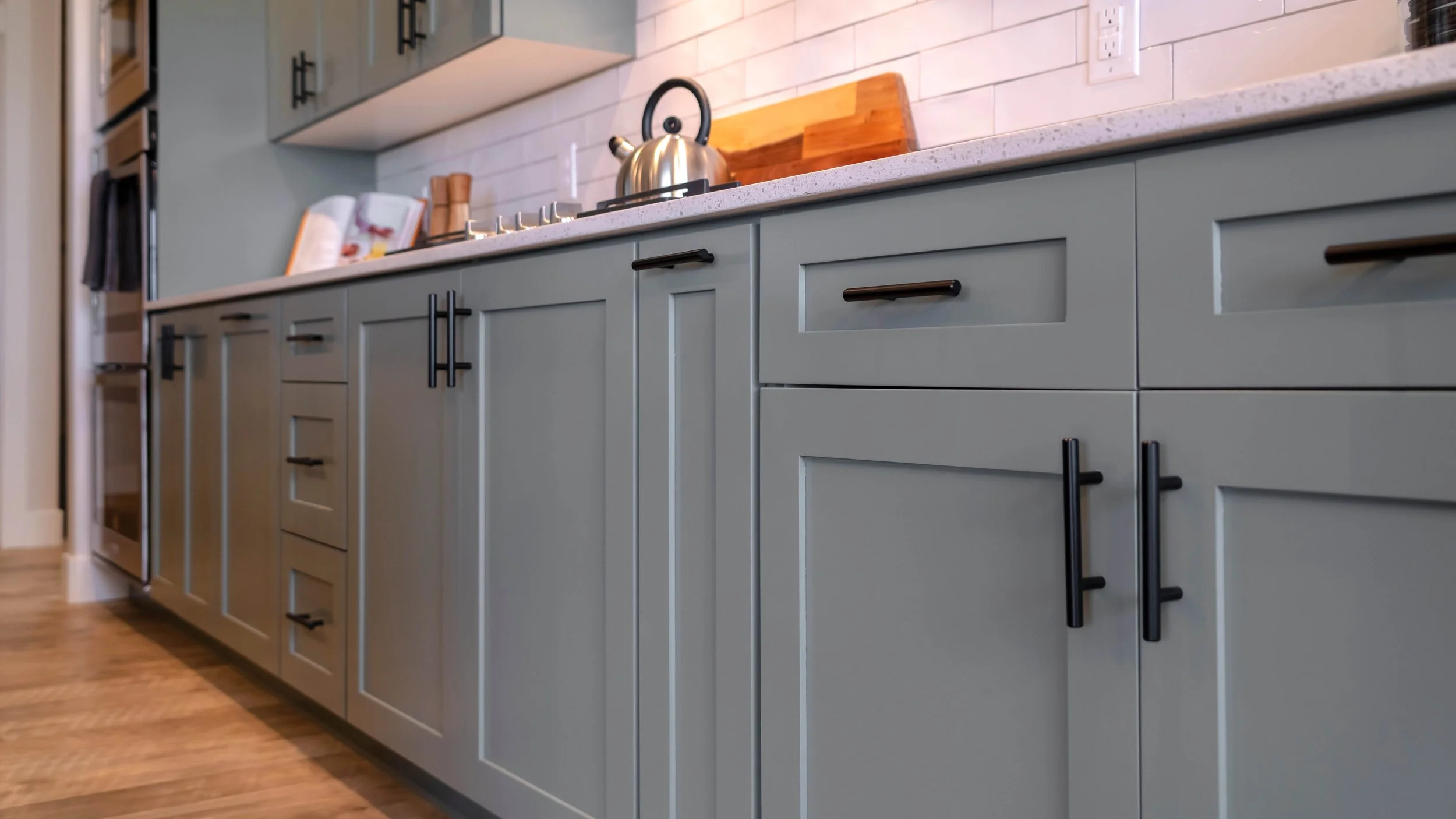 View of light gray kitchen cabinets with black handles, a white marble countertop, and a white brick backsplash. There is a stainless steel kettle on the stove and a cutting board behind it.