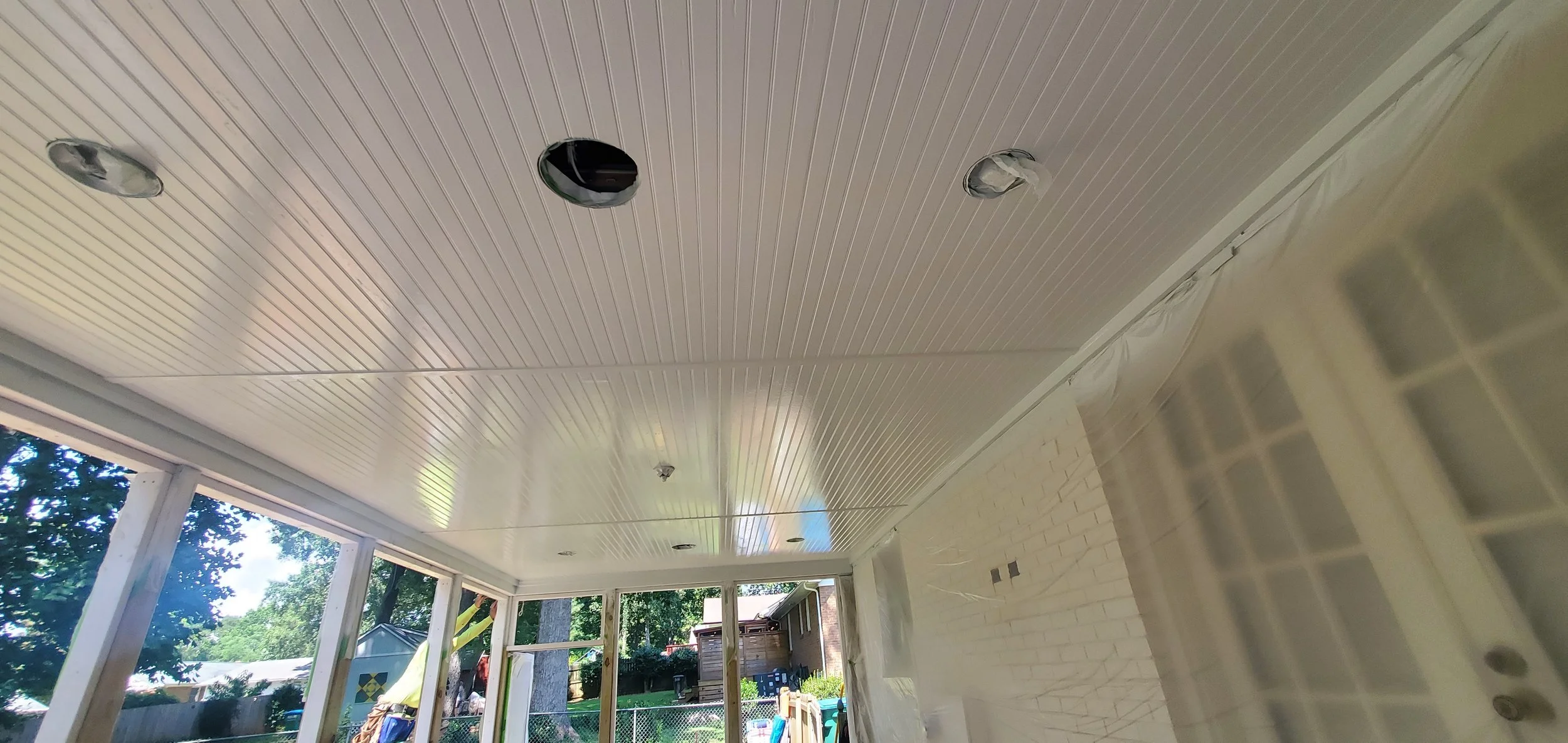 The ceiling of a porch with white beadboard paneling and several light fixtures, some of which are missing covers, and a view of the outside yard with trees, a shed, and a chain-link fence.