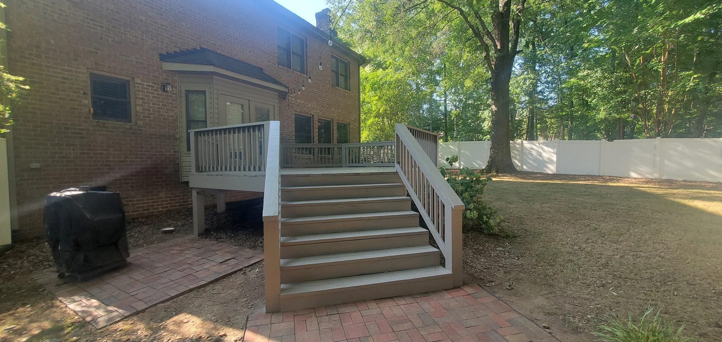 Backyard with wooden stairs leading to a deck attached to a brick house, surrounded by trees, with a white fence in the background.