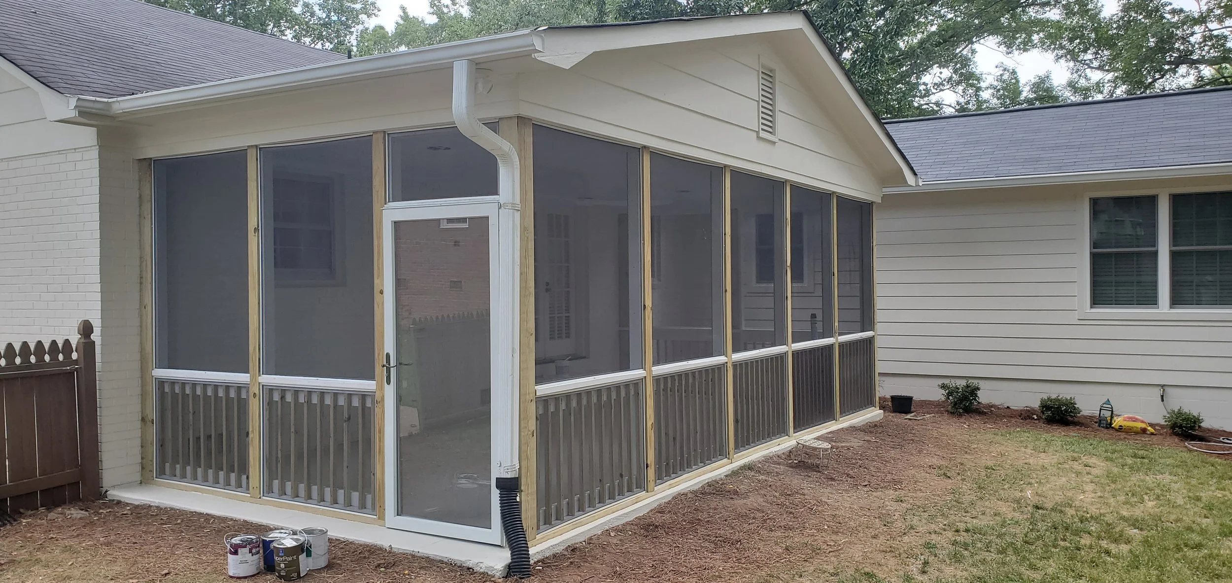 Unfinished screened porch attached to a house with white siding, construction materials, and a small garden area with bushes.