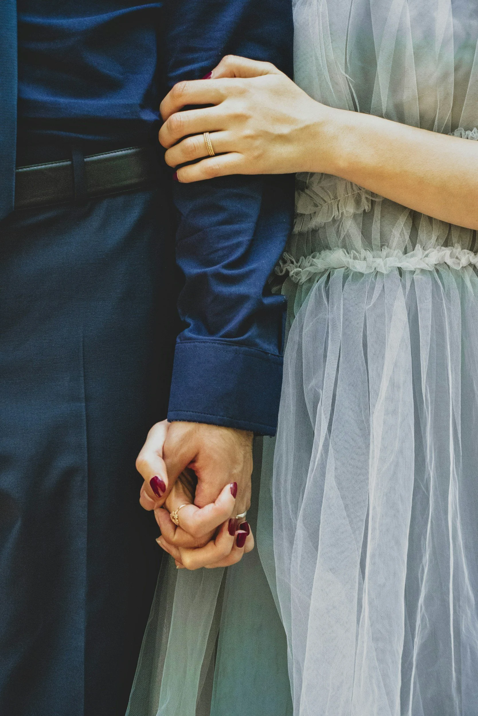 Close-up of a couple holding hands, with the woman's hand resting on the man's arm. The woman is wearing a light-colored, sheer, ruffled dress and a gold ring on her finger, while the man is dressed in dark clothing with a black belt.