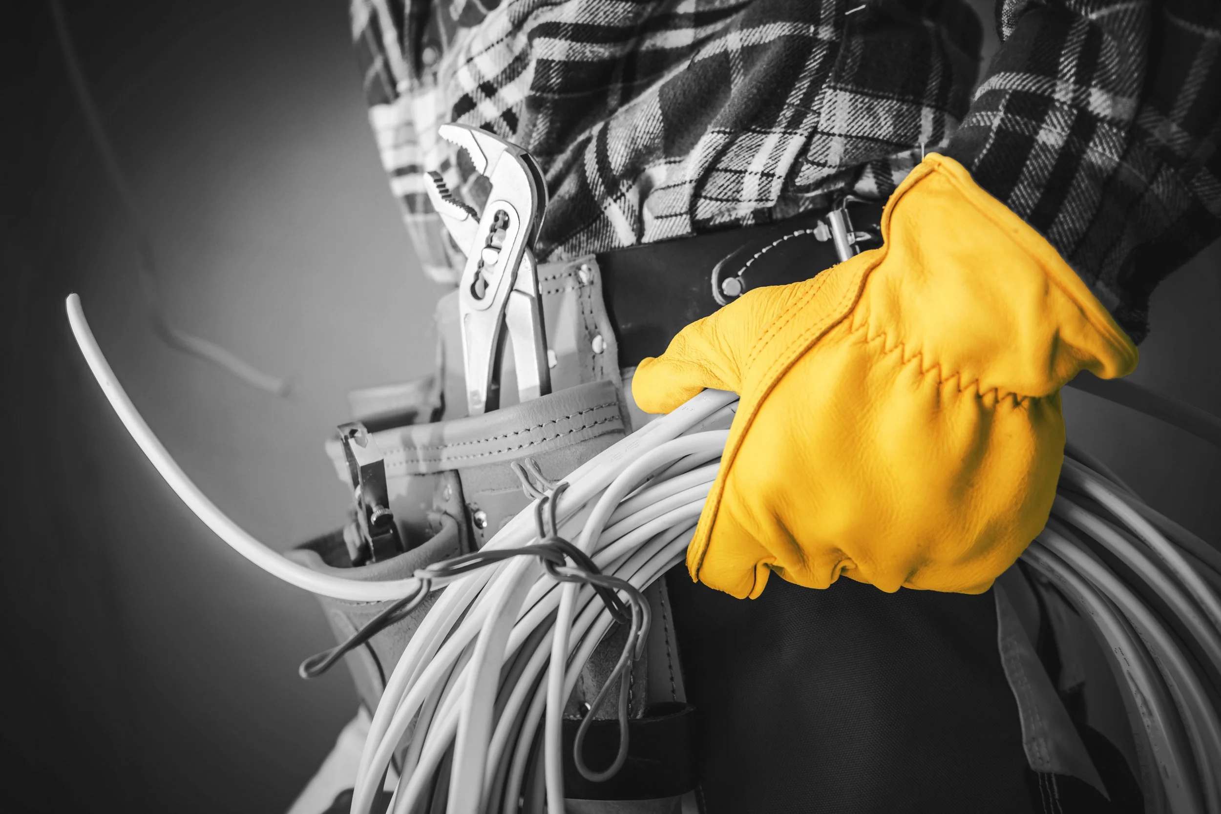 Close-up of an electrician's tool belt with electrical wires, pliers, and a wrench, with yellow gloves and plaid shirt visible, in black-and-white with the gloves in color.