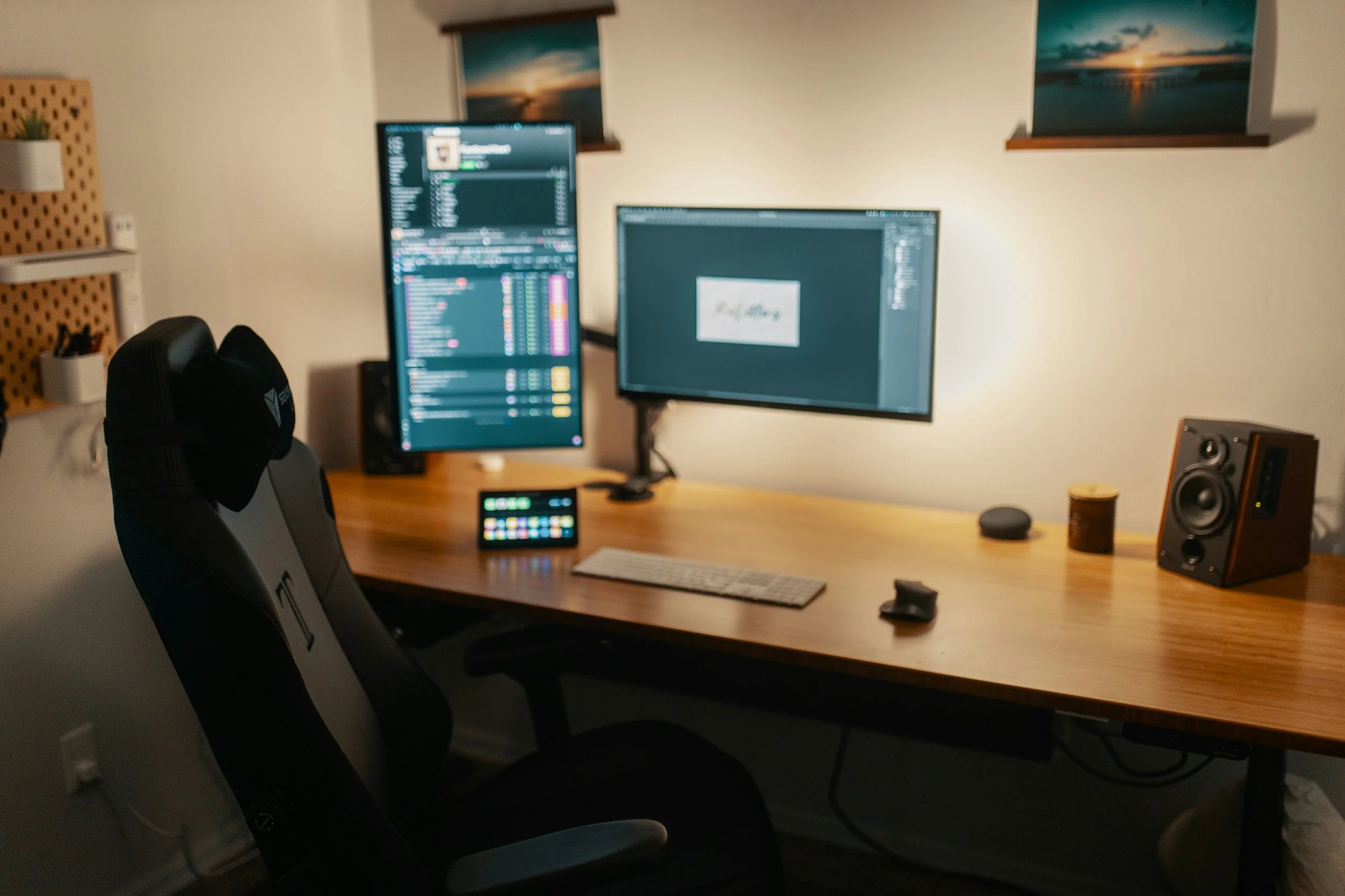 A workspace with a wooden desk holding two computer monitors, one displaying a messaging app and the other a graphics editing program. There are two ergonomic chairs in front of the desk, and various small items like a keyboard, mouse, speaker, and a container on the desk. Behind the workspace, two windows with sunset or sunrise views are visible.