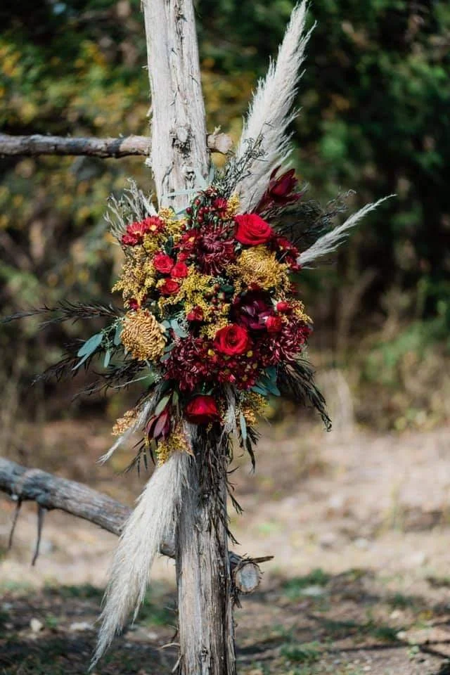A floral arrangement with red roses, yellow, and dark red flowers attached to a vertical tree branch outdoors.