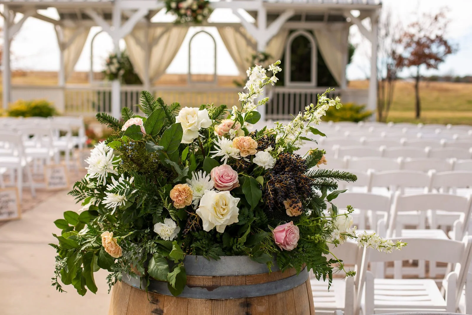 A large floral arrangement with white, pink, and peach flowers and green foliage, placed on a wooden barrel at an outdoor wedding ceremony setup with white chairs and a decorated gazebo in the background.
