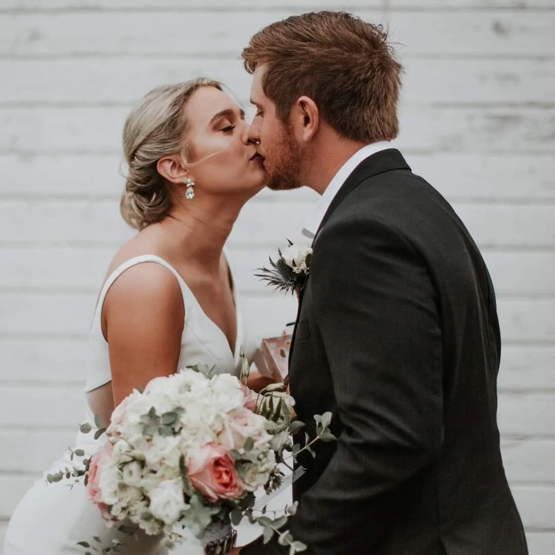 A bride and groom sharing a kiss on their wedding day, with the bride holding a bouquet of white and pink flowers, and the groom dressed in a black tuxedo. There is a white wooden background.
