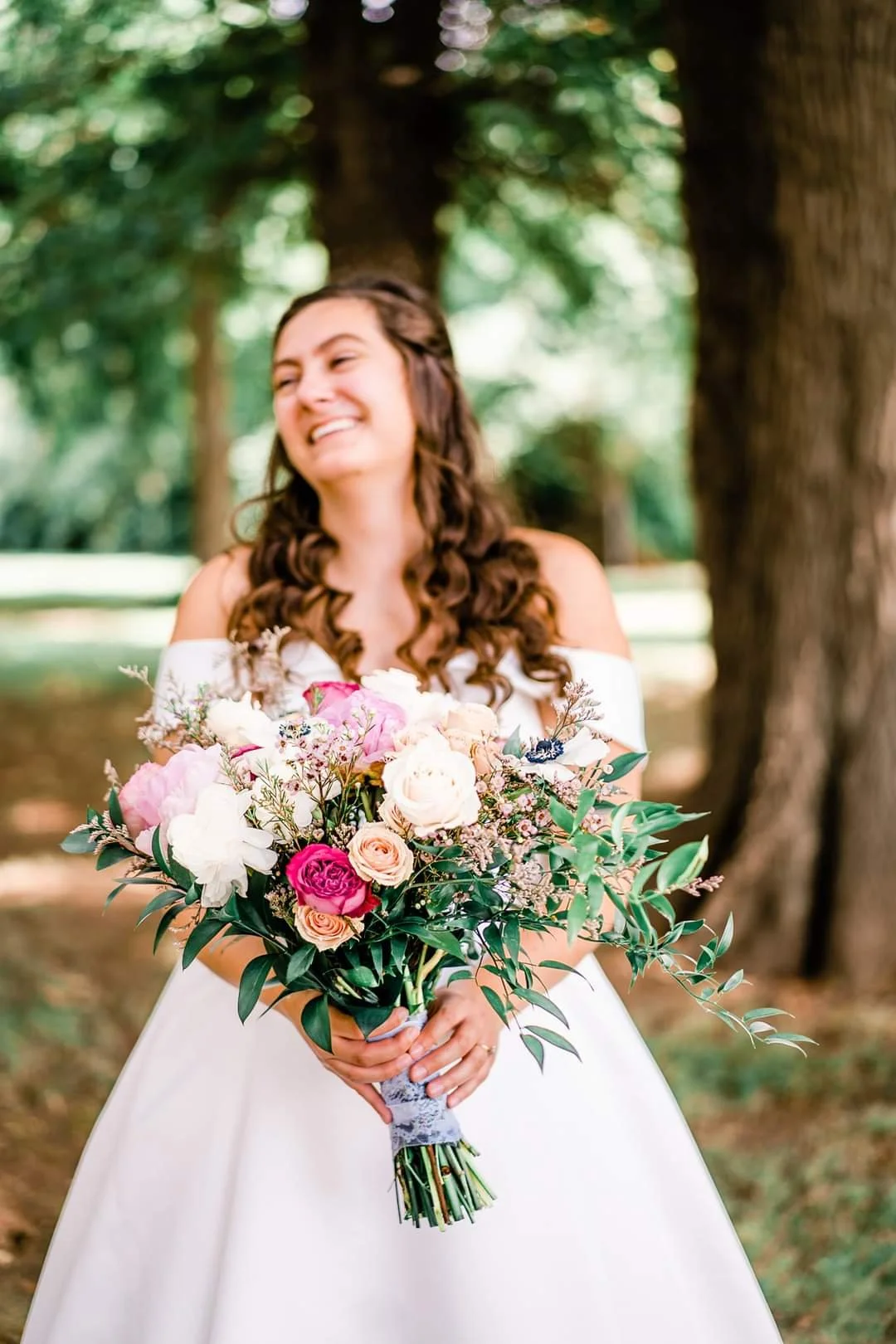 A smiling woman in a white dress holding a large bouquet of pink, white, and cream flowers, standing outdoors near trees.