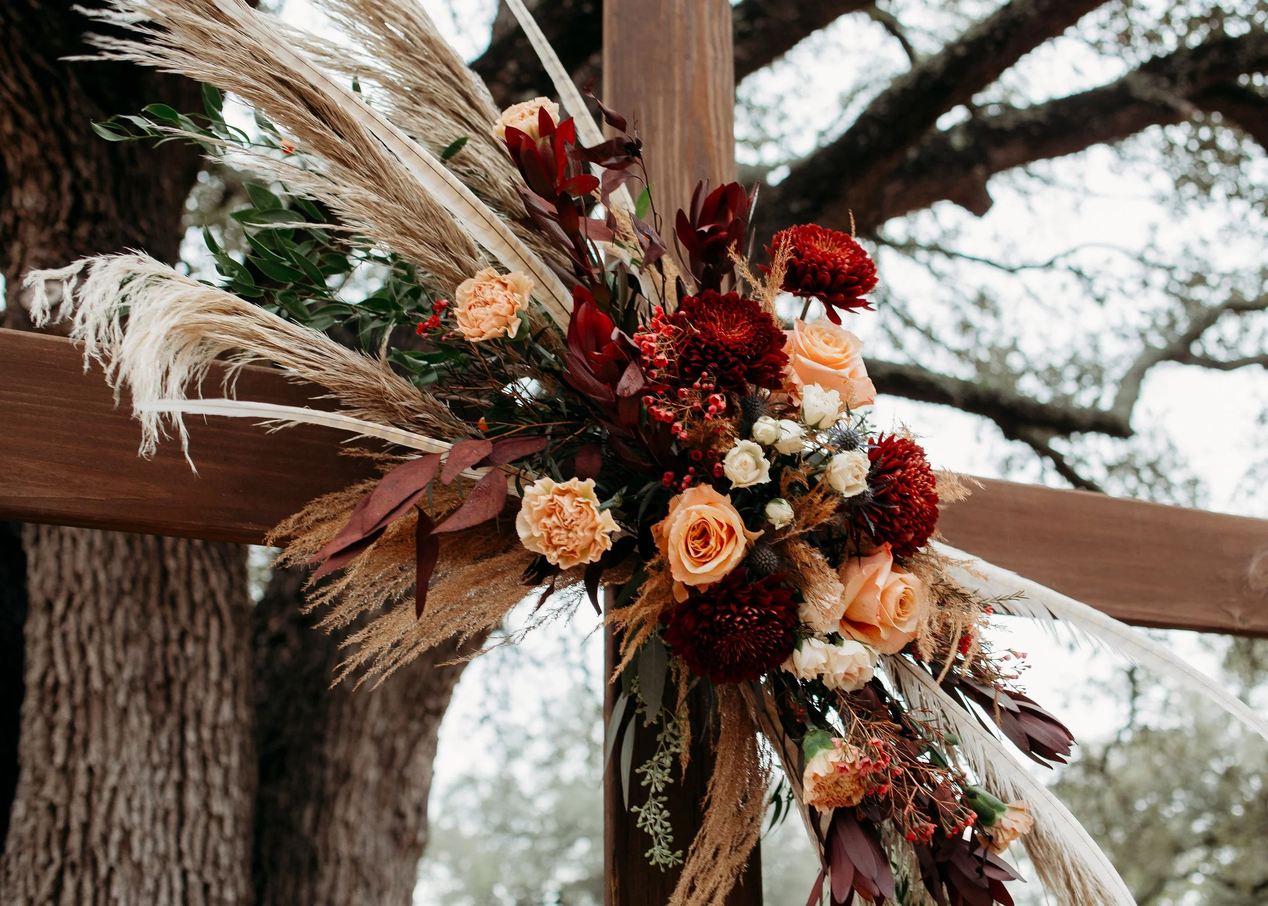 Decorative floral arrangement with peach and red flowers attached to a wooden structure outdoors, with large tree and cloudy sky in background.
