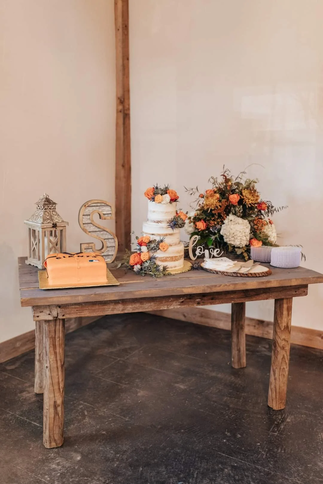 Wooden table decorated with floral wedding cake, bouquet, lantern, and love sign, set against plain wall.