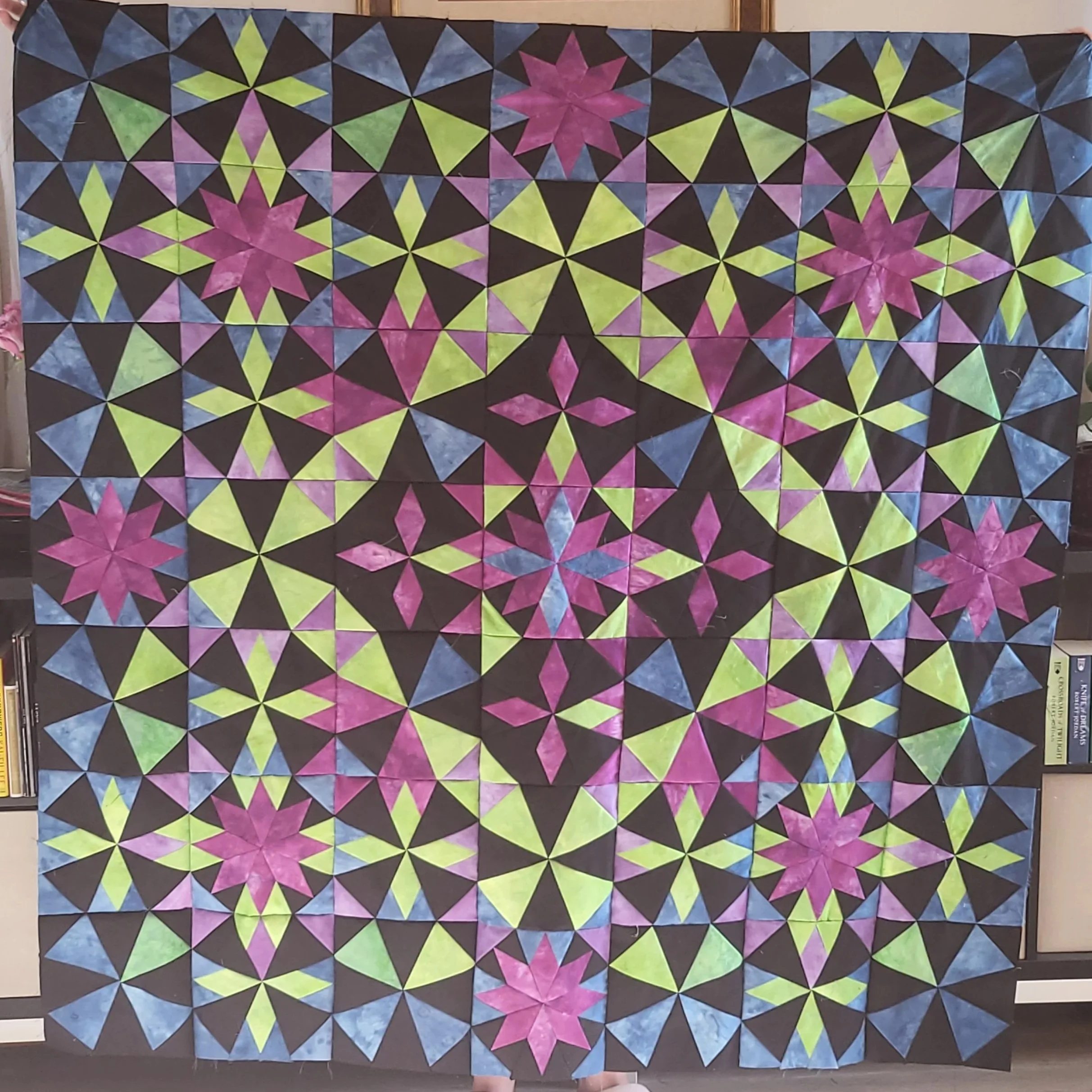 Colorful quilt with geometric star patterns in pink, green, blue, and black, displayed on a wall.