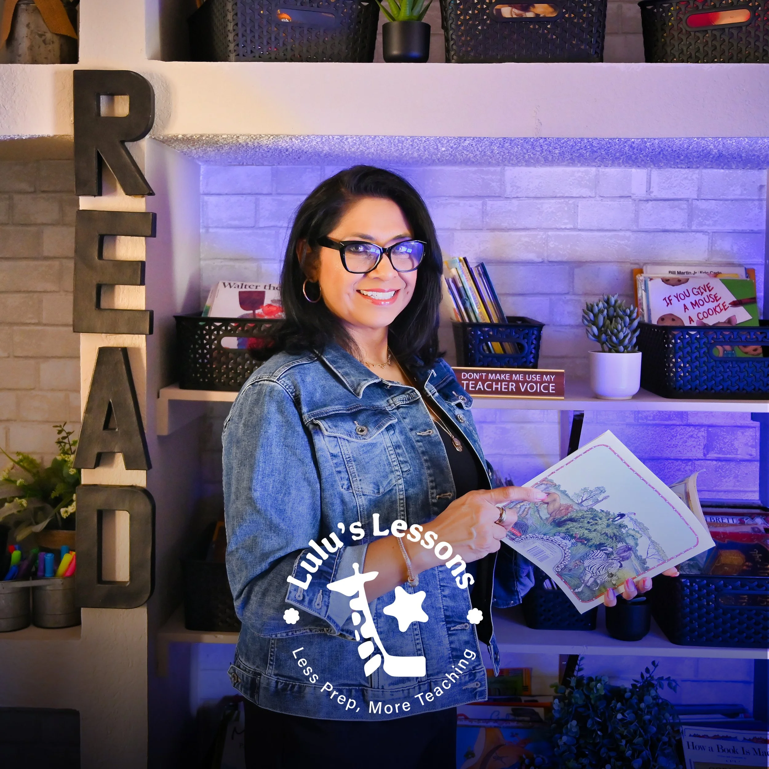 A woman with black hair and glasses smiling while holding an open book in a room decorated with books, plants, and decorative items. There is a large black letter "R" on the wall to her left, and various books and decorative items on shelves behind her.