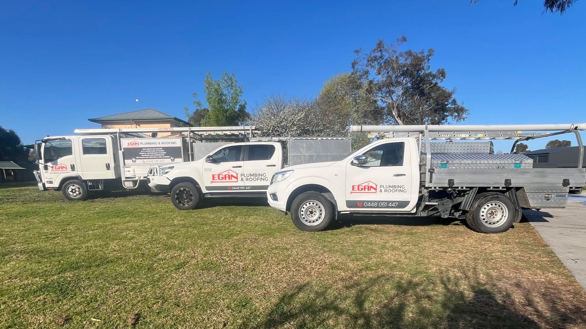 Three Egan Plumbing & Roofing trucks parked on grass with trees and a house in the background under a clear blue sky.