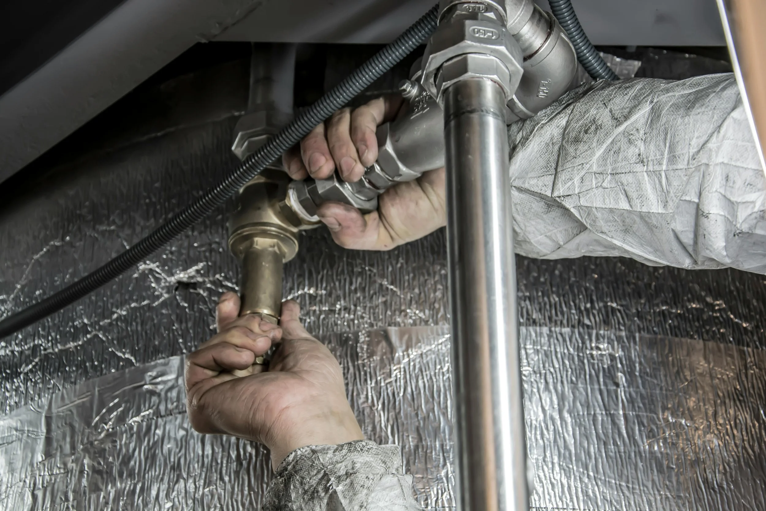 Two hands, one holding a wrench, working on a pipe connection under a vehicle. The area around is insulated with reflective material and covered with a grayish sheet.