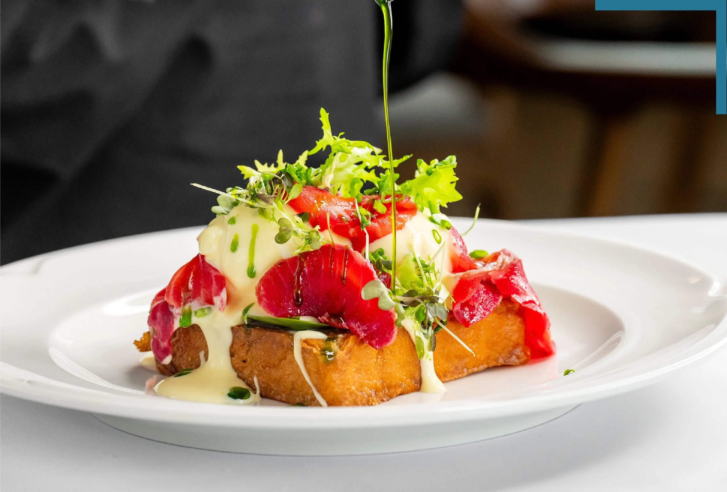 Close-up of an elegant dish with greens, tomatoes, microgreens, and a drizzle of sauce on a white plate, with a blurred background.