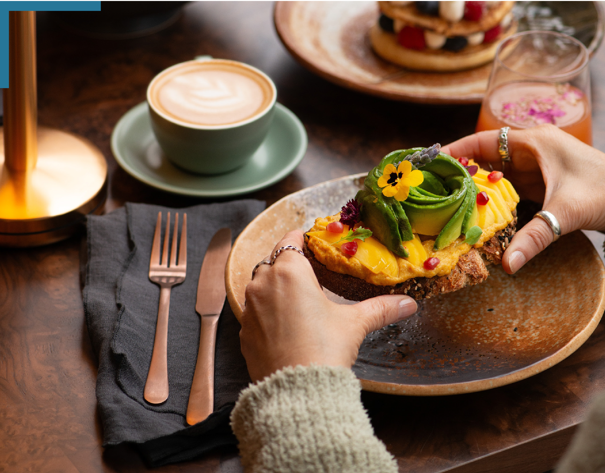 Hands holding a slice of avocado toast topped with edible flowers, pomegranate seeds, and herbs on a wood table with coffee, water, and a fruit tart in the background.