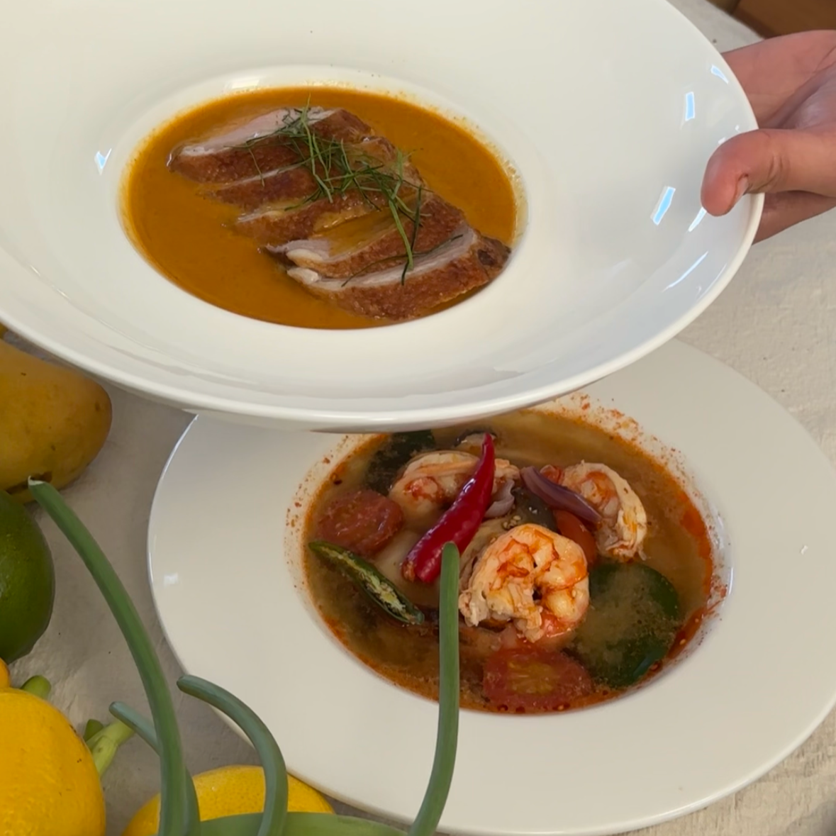 A white bowl holding a serving of red curry with shrimp, tomato, onion, and chili pepper. Nearby, a larger plate contains red curry with sliced fish, shrimp, and various vegetables, garnished with herbs. Lemons are visible in the background.