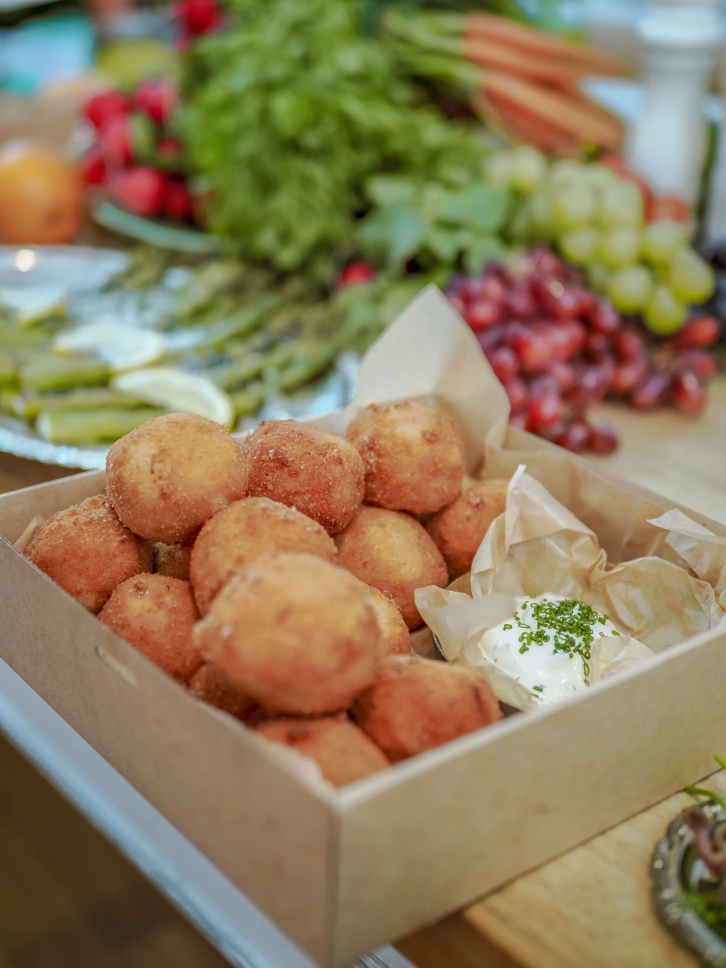 Basket of fried bites with a side of dipping sauce and chopped herbs, with a colorful array of fruits and vegetables in the background.