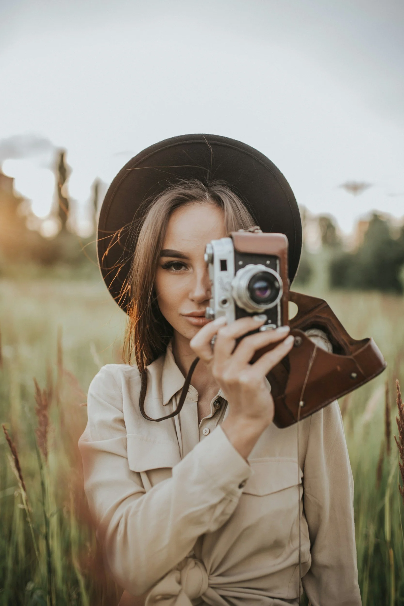 A young woman wearing a wide-brimmed hat and beige jacket taking a photo with a vintage camera in a field of tall grass.