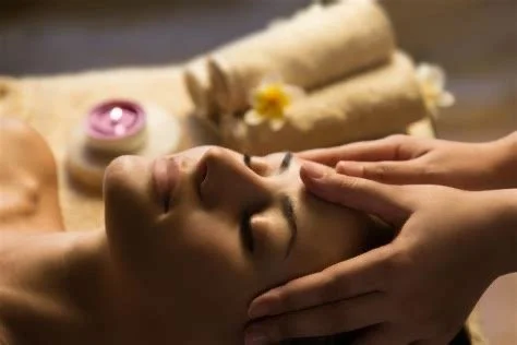 Person receiving a facial massage at a spa with rolled towels, candles, and flowers in the background.