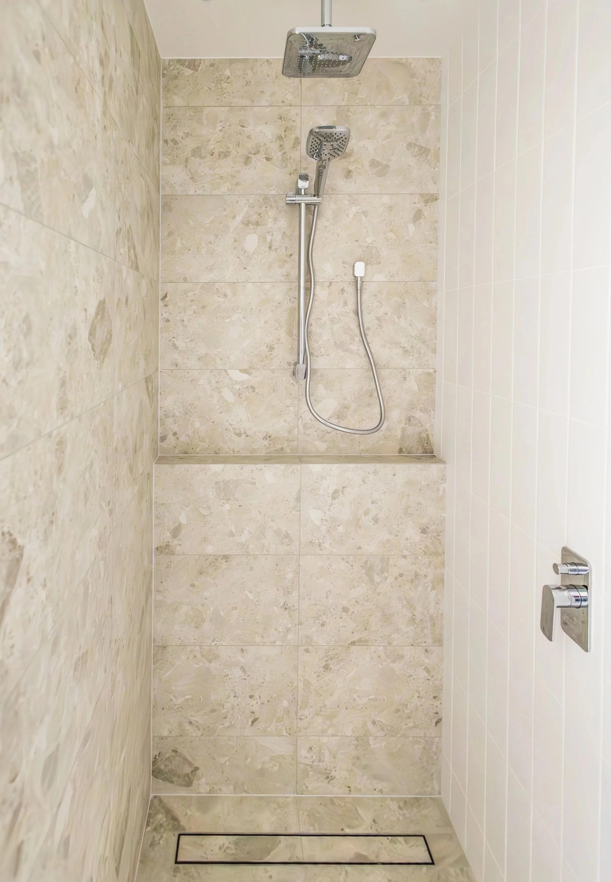 A modern walk-in shower with beige marble tiles on the walls and floor, a rainfall showerhead, and a handheld shower attachment, all enclosed by a glass door.
