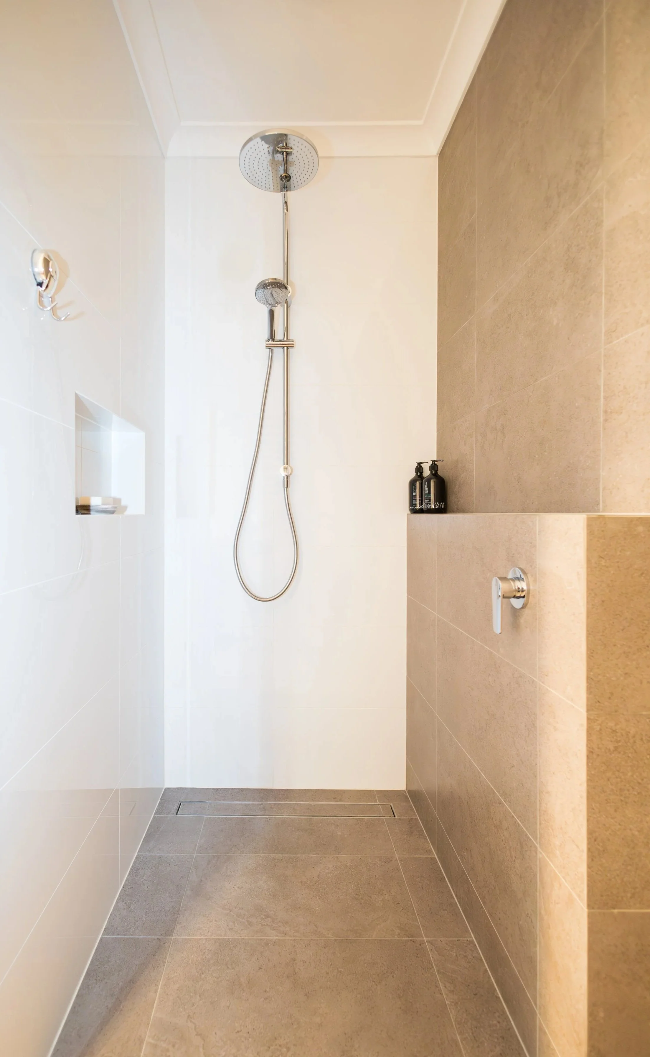 Modern walk-in shower with a rainfall showerhead, a handheld shower, and a small built-in shelf with soap, with beige and white tiles.