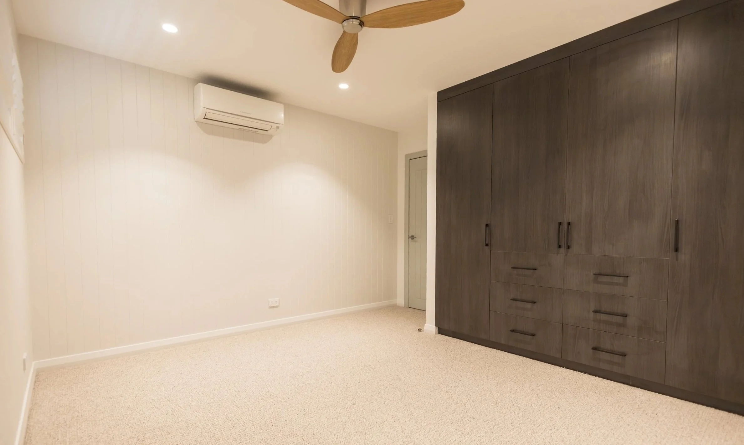 Room with beige carpet, white wall with vertical paneling, white air conditioning unit, ceiling fan, closed grey door, and large dark brown built-in wardrobe with black handles.