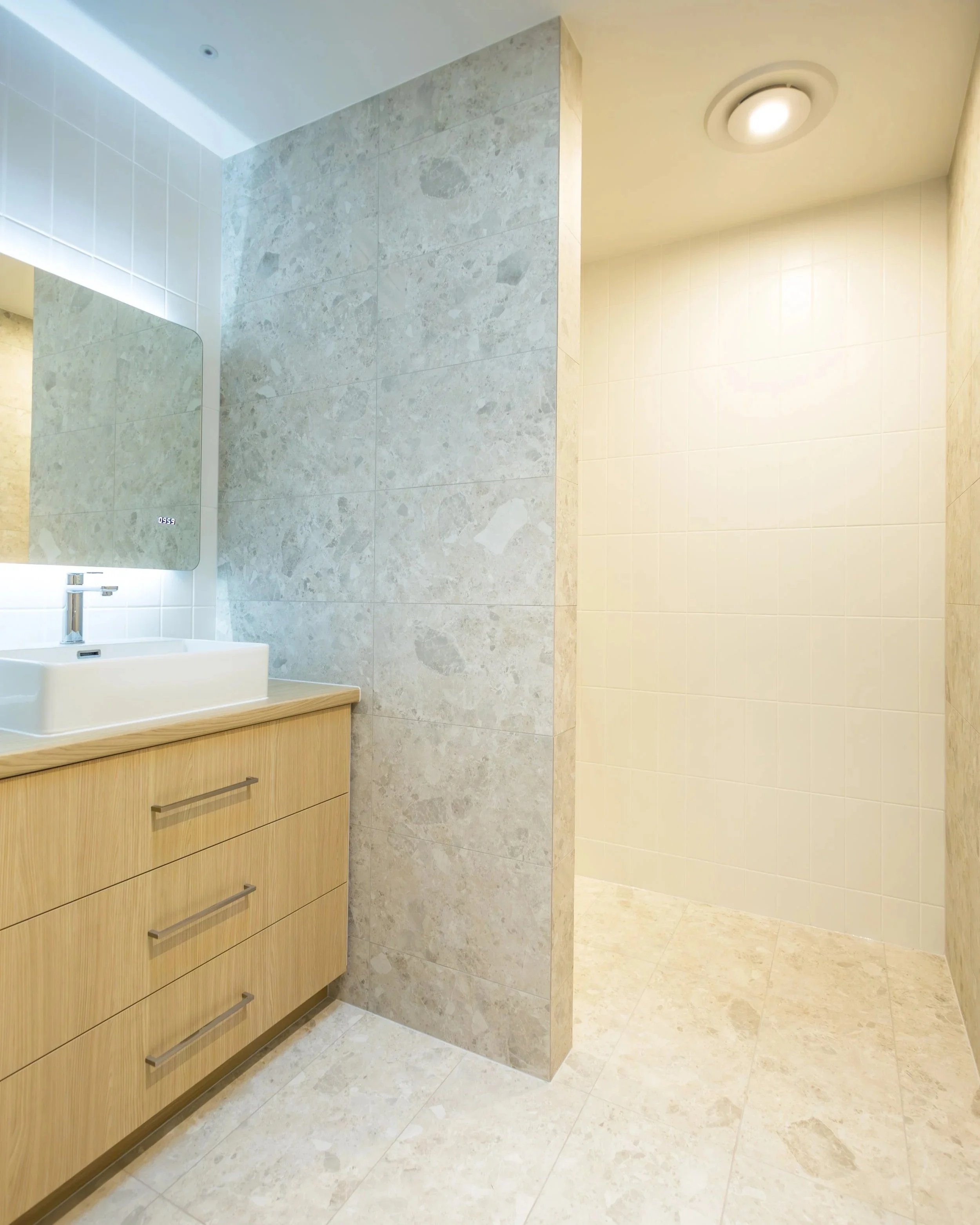 Modern bathroom with light wood vanity, white sink, large mirror, tiled walls, and a walk-in shower with a ceiling light.