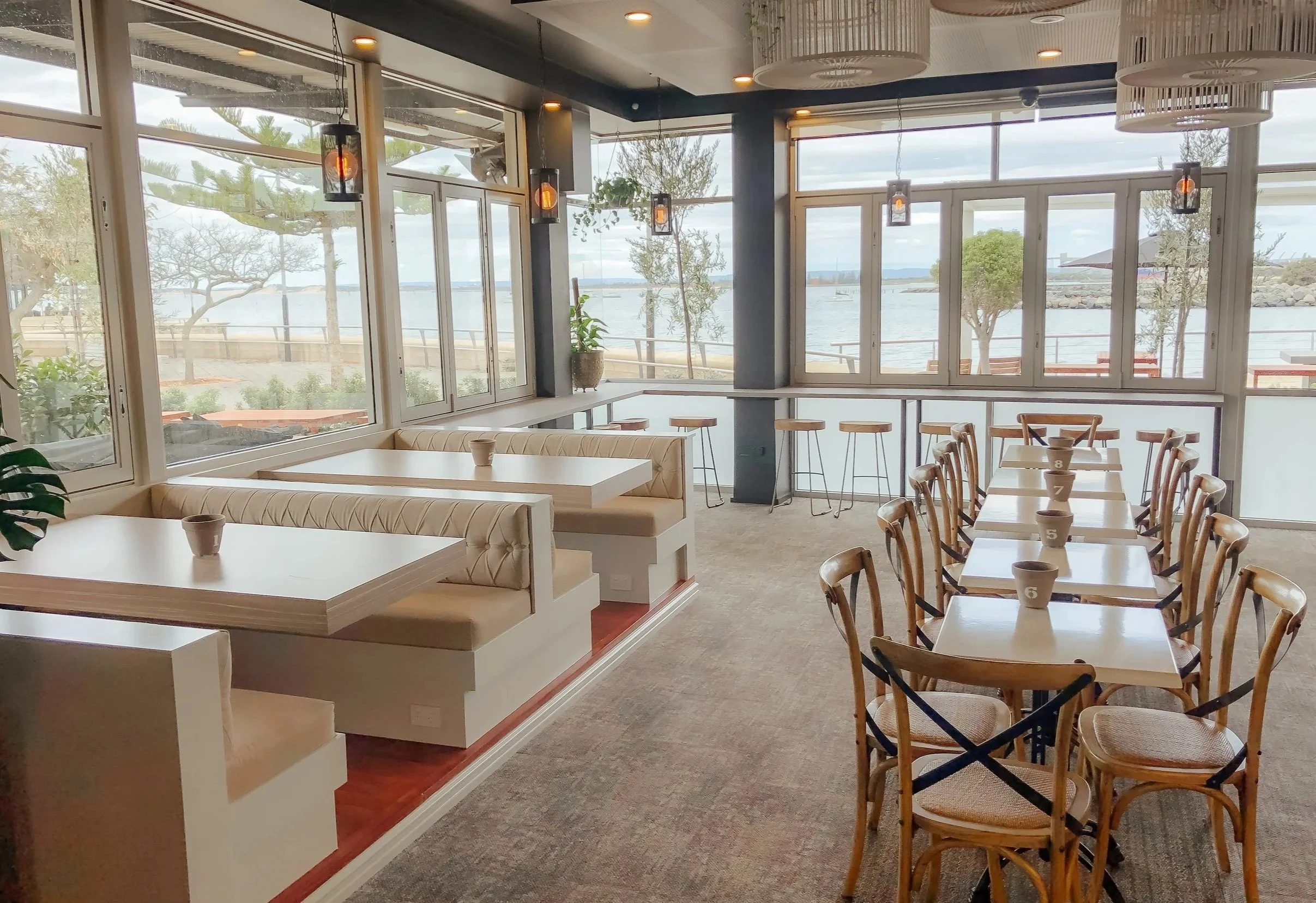 Interior of a bright dining area overlooking a waterfront with large windows, featuring tables and chairs, some numbered, and a seating area with booths and bar stools.