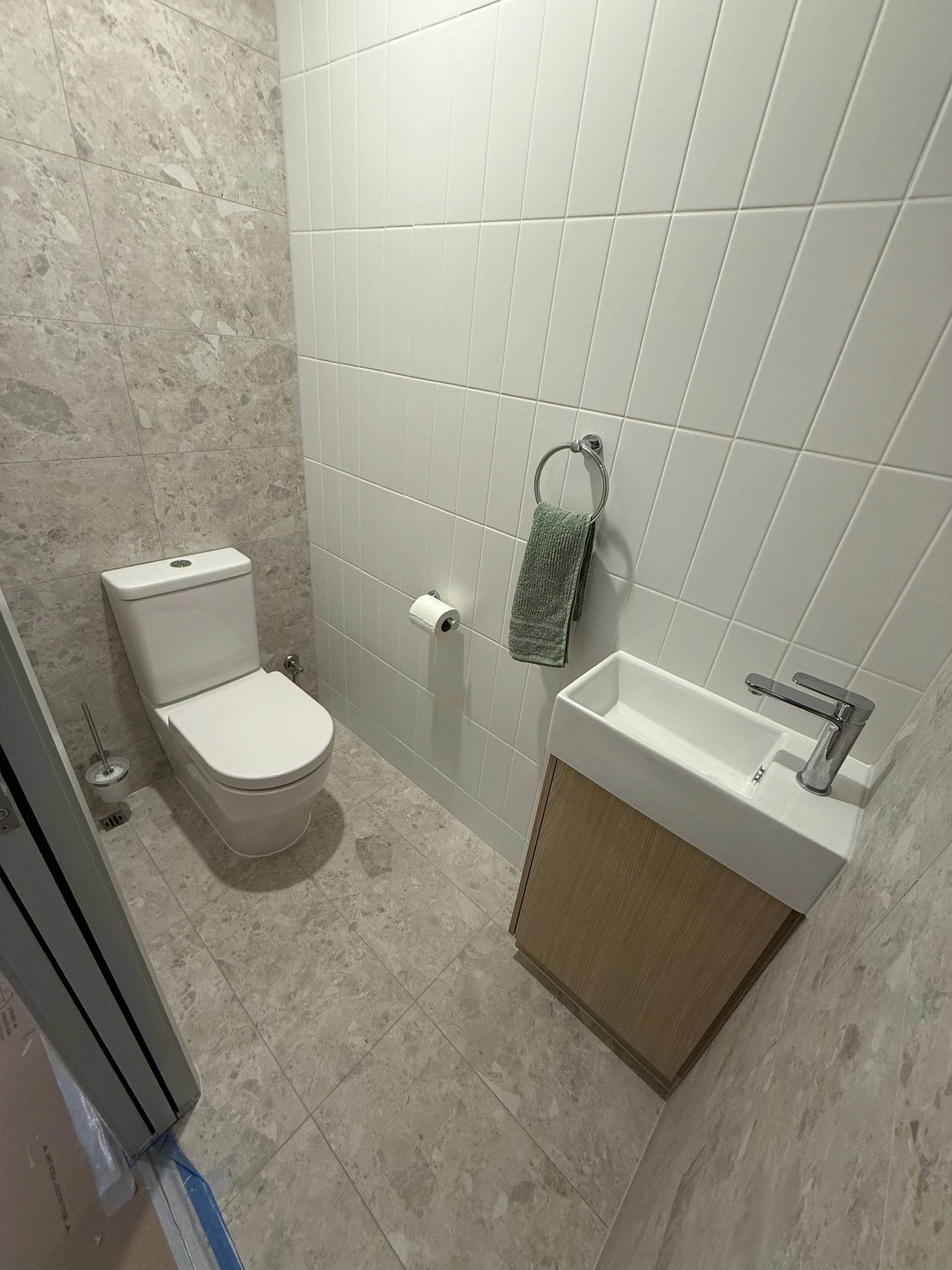 Small modern bathroom with a white toilet, a small sink with a wooden cabinet, a toilet paper roll, a green towel on a ring, and a beige tiled floor and wall with partial wall tiles.