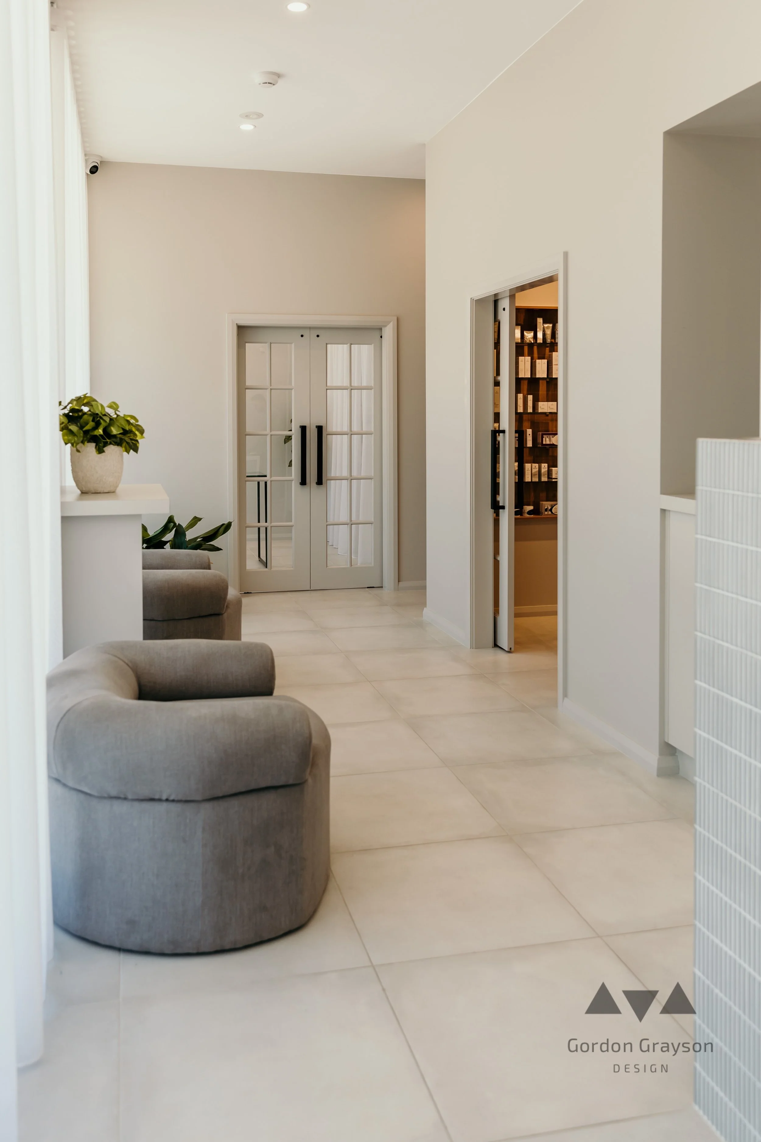 Modern interior with beige upholstered chairs, potted plants, white walls, light-colored tiled floor, and double glass-paneled doors. Aesthetic commercial builds western australia