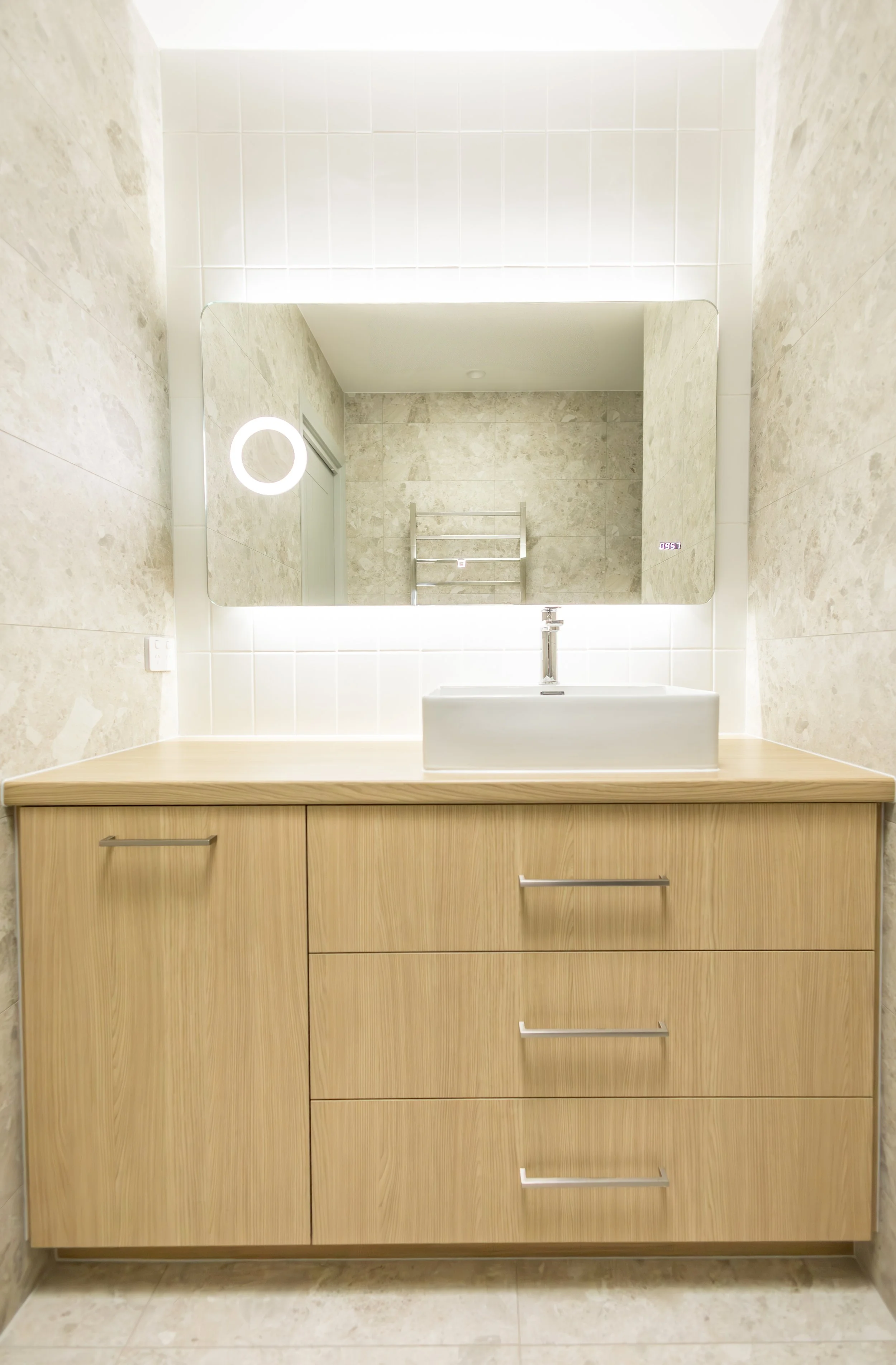 Bathroom vanity with a rectangular vessel sink, large mirror, and wood cabinet drawers, with neutral-colored tiled walls and floor.
