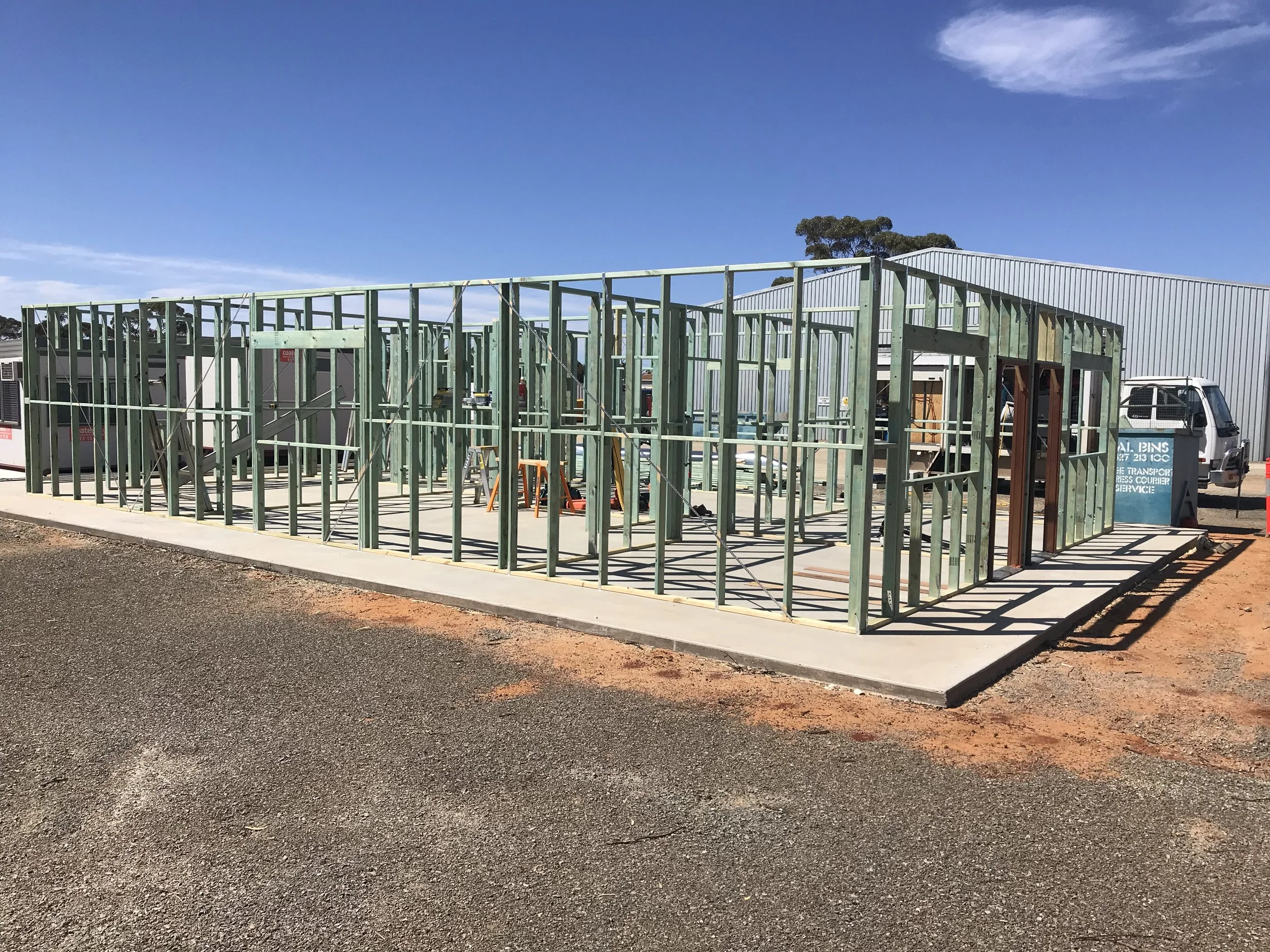 Construction site of a commerical build with green wooden framing, on a concrete slab, under a clear blue sky.