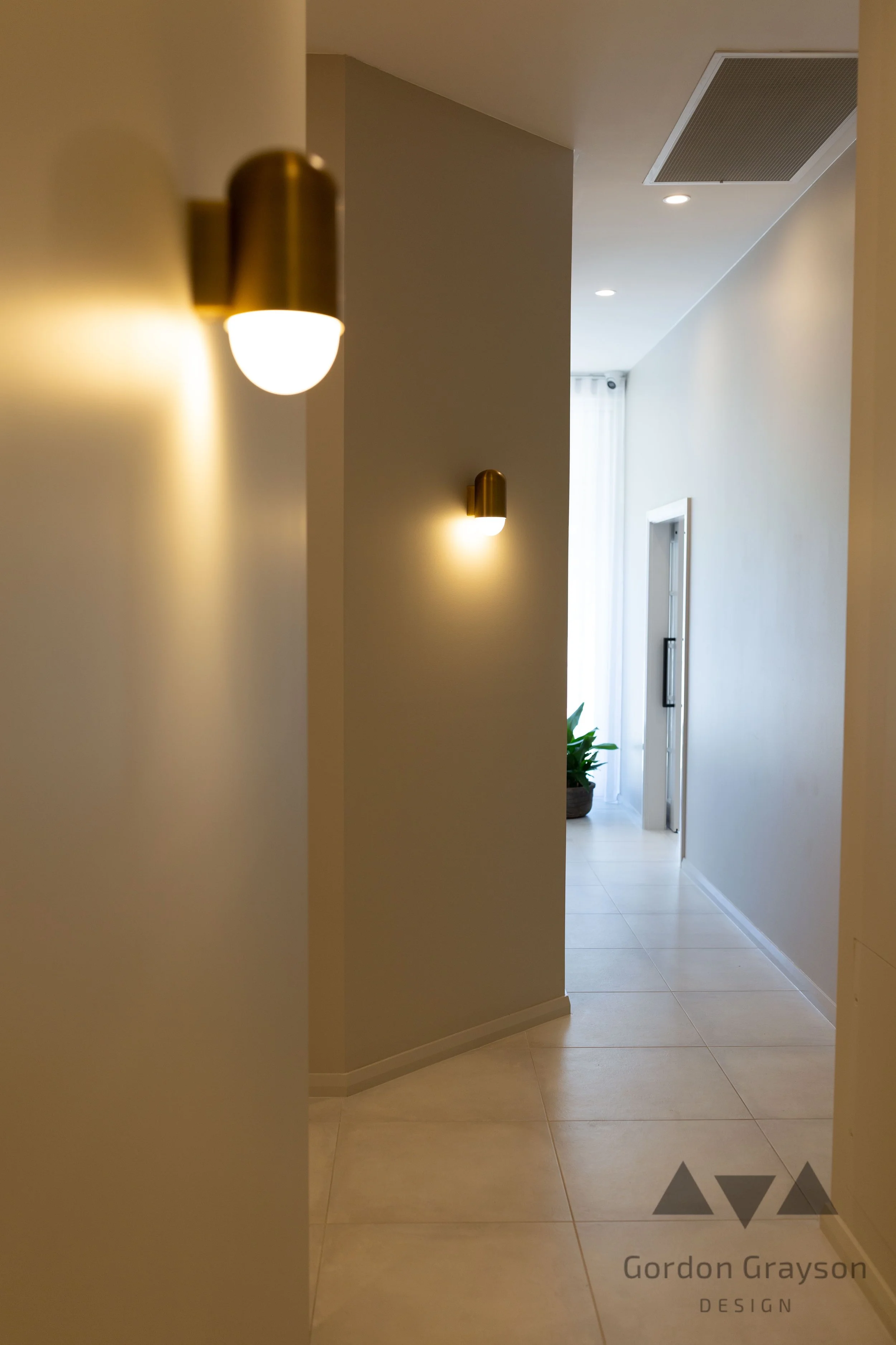 Interior hallway with beige tiles, wall-mounted gold light fixtures, a potted plant near a window with sheer curtains, and a door at the end of the hallway.