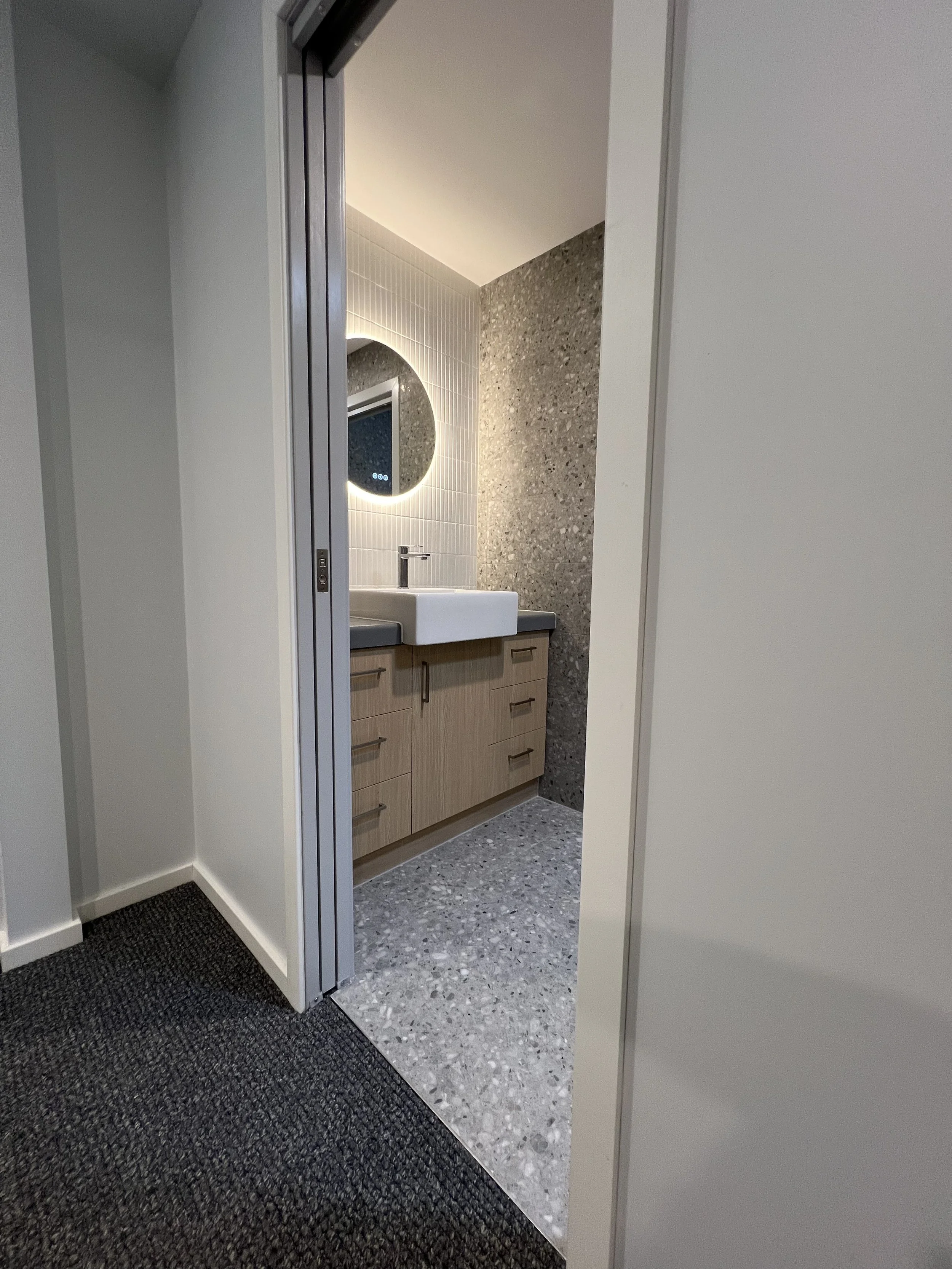 View of a modern bathroom with a white sink, wood vanity, round backlit mirror, and textured gray wall, seen through an open folding door from an adjacent room with dark carpet.