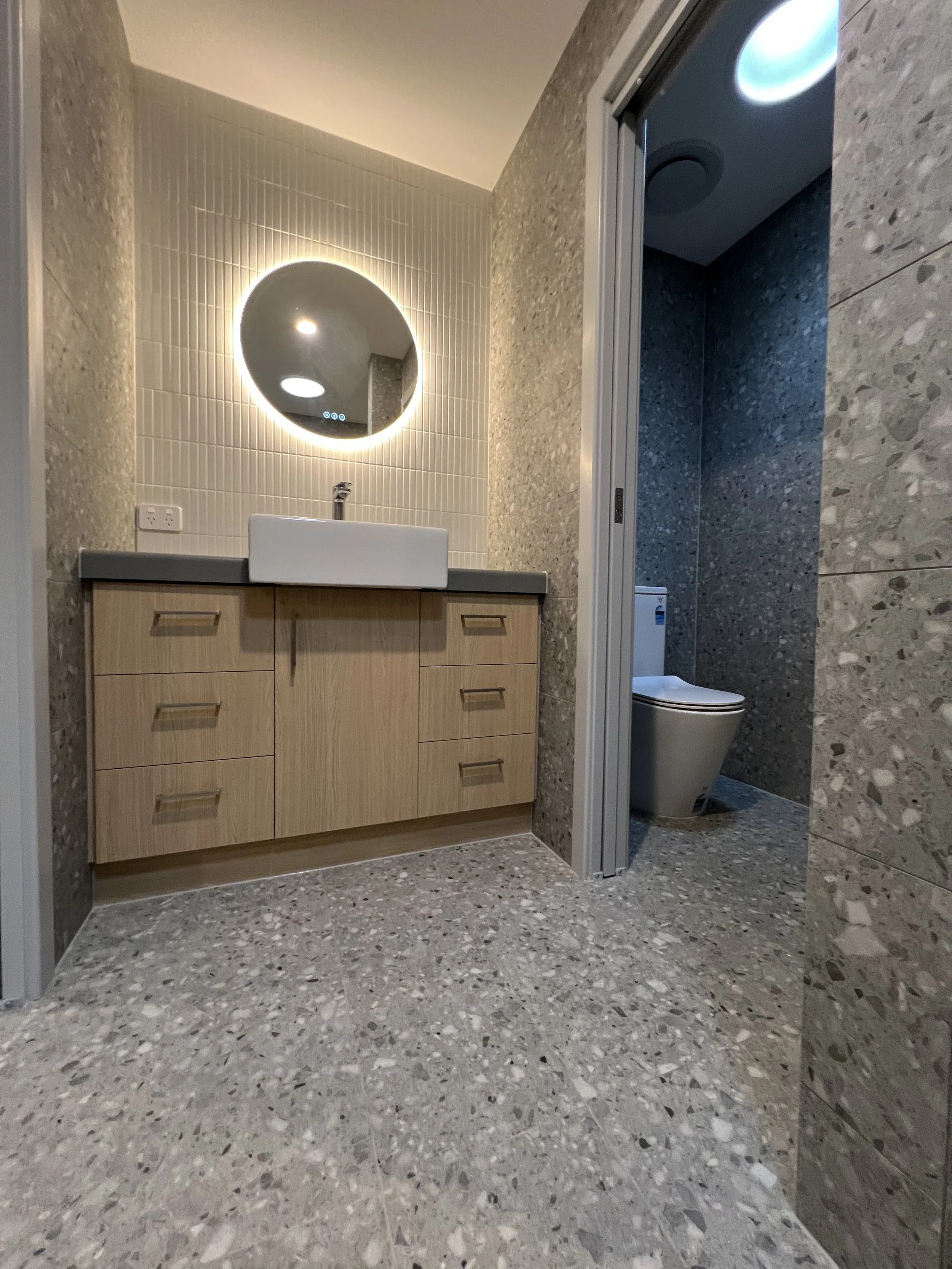 Modern bathroom renovation with wooden vanity, white sink, illuminated round mirror, and a separate toilet area.