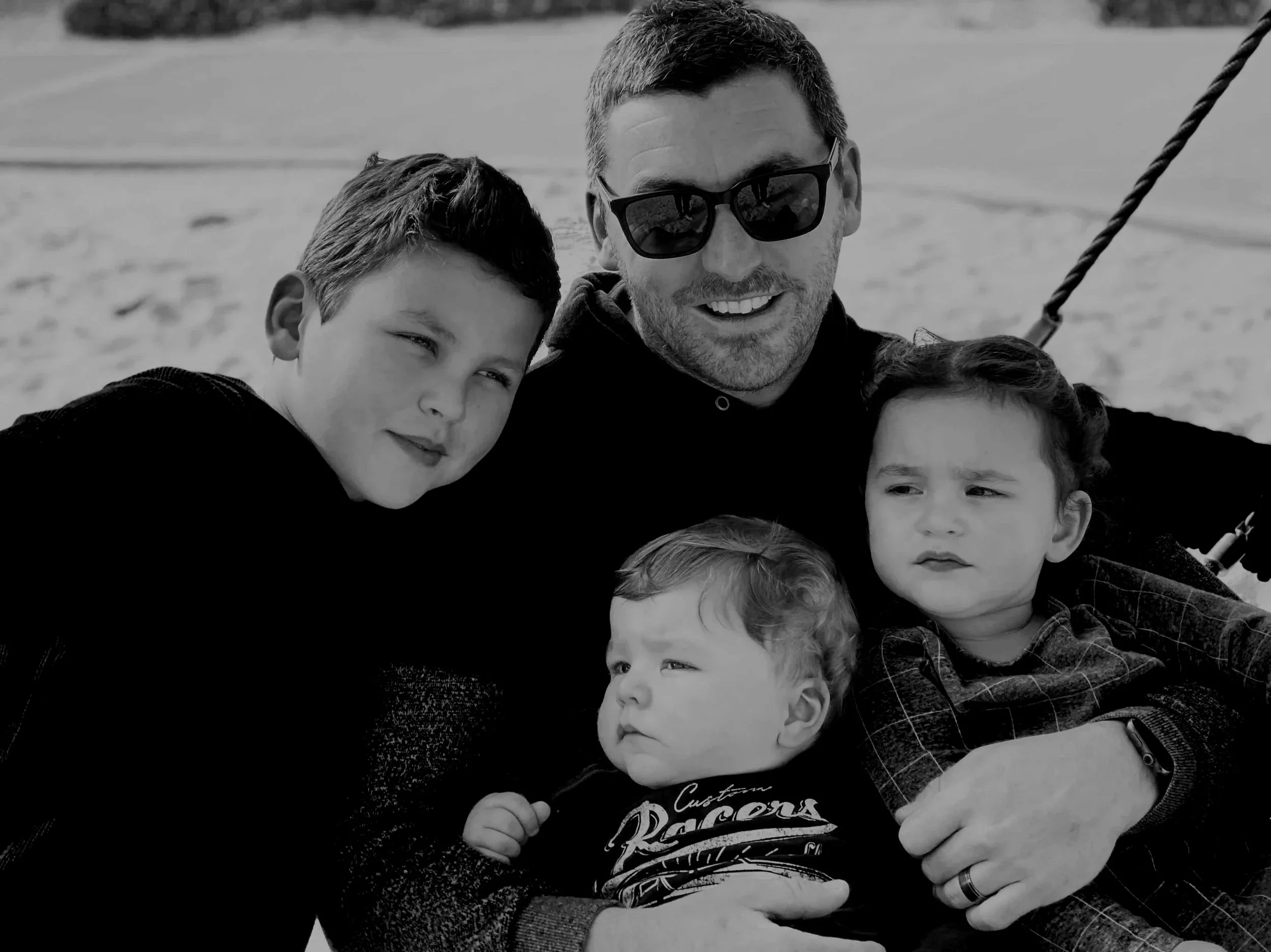A man with sunglasses and three children sitting on a swing at the beach.