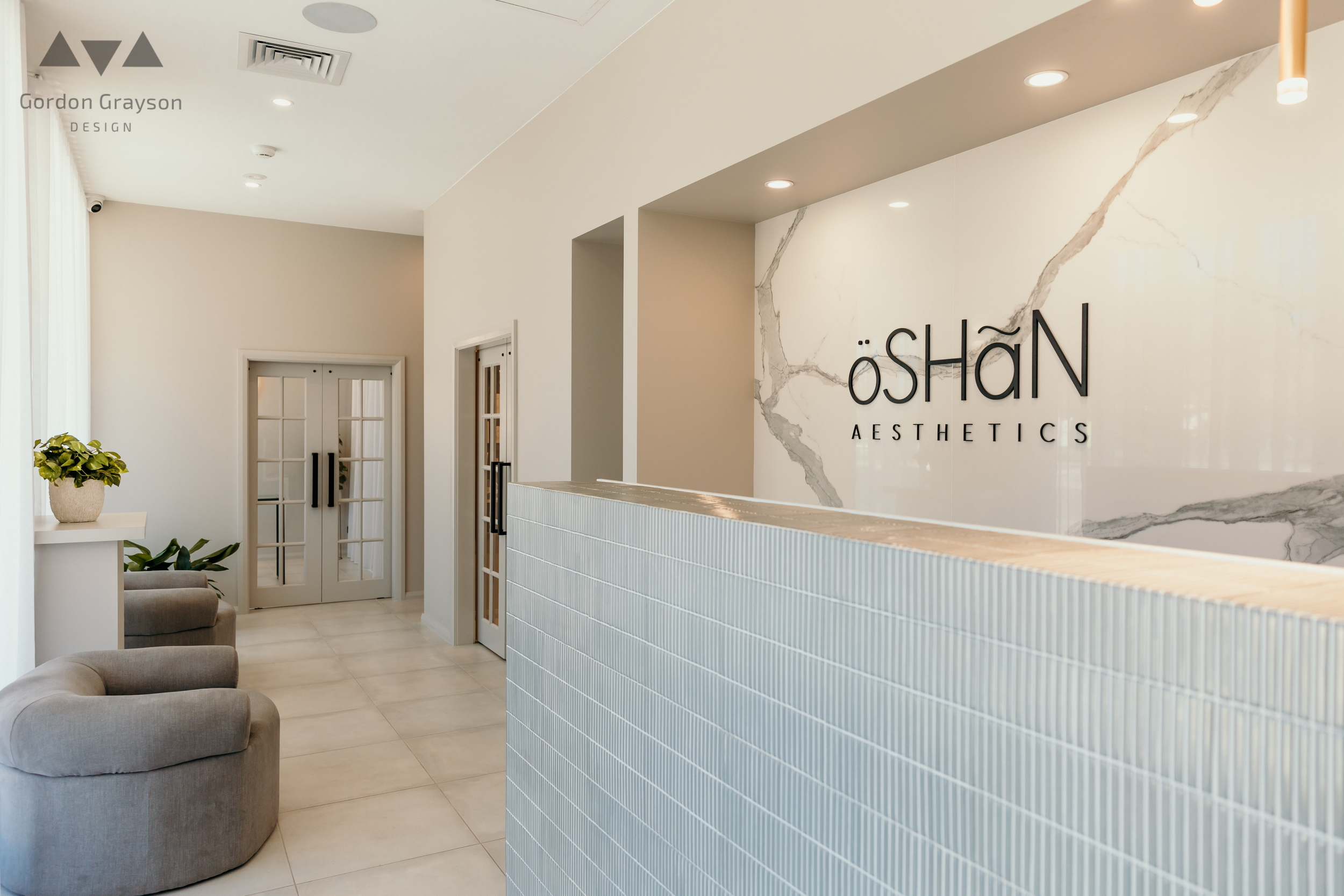 Interior of a modern aesthetics clinic with a reception desk, the clinic's logo 'Öshan Aesthetics' on a marble wall, and waiting area with gray chairs and potted plants.