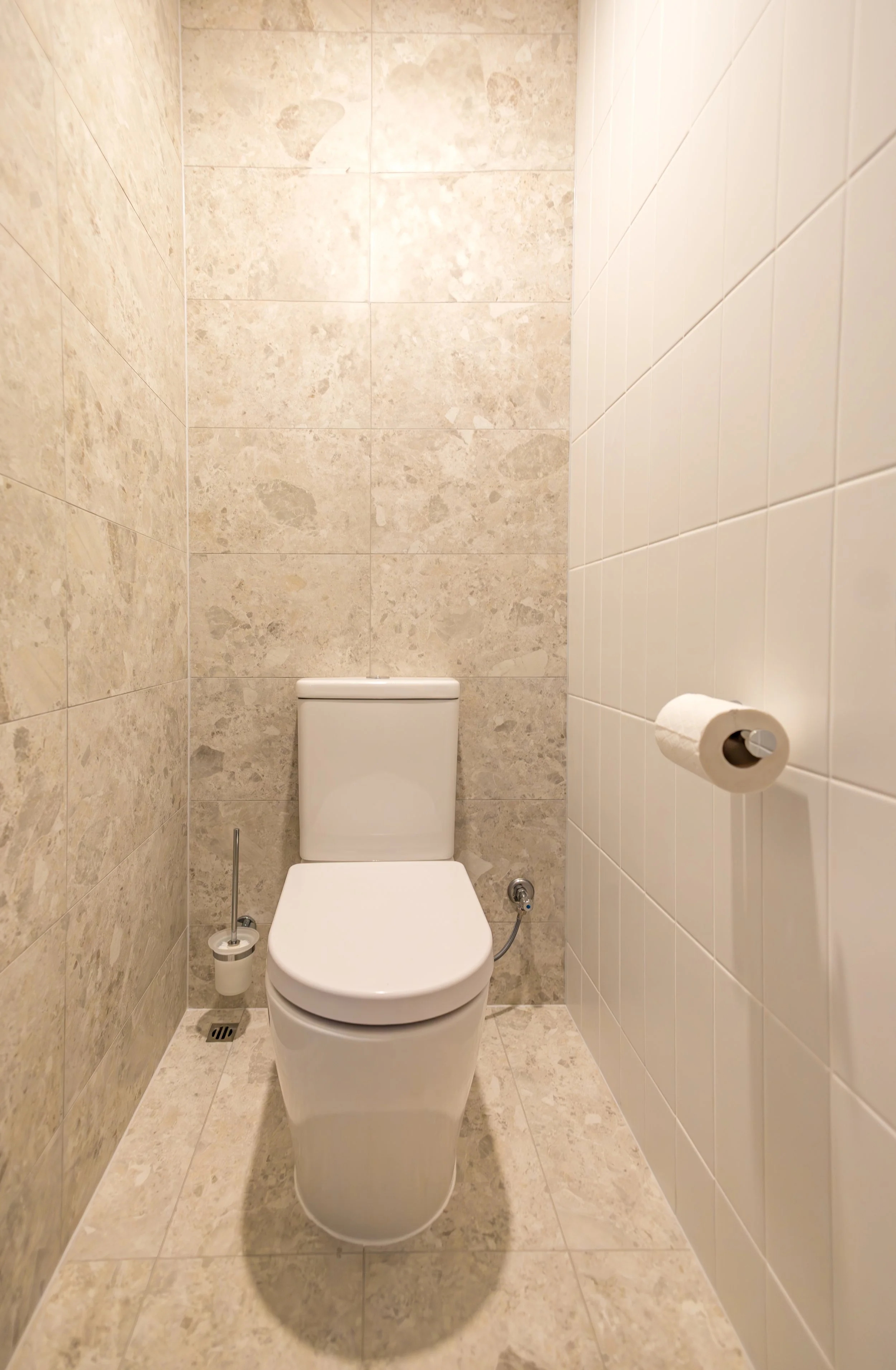 A small bathroom with beige tiled walls and floor, featuring a white toilet, in a custom granny flat built by busselton builders