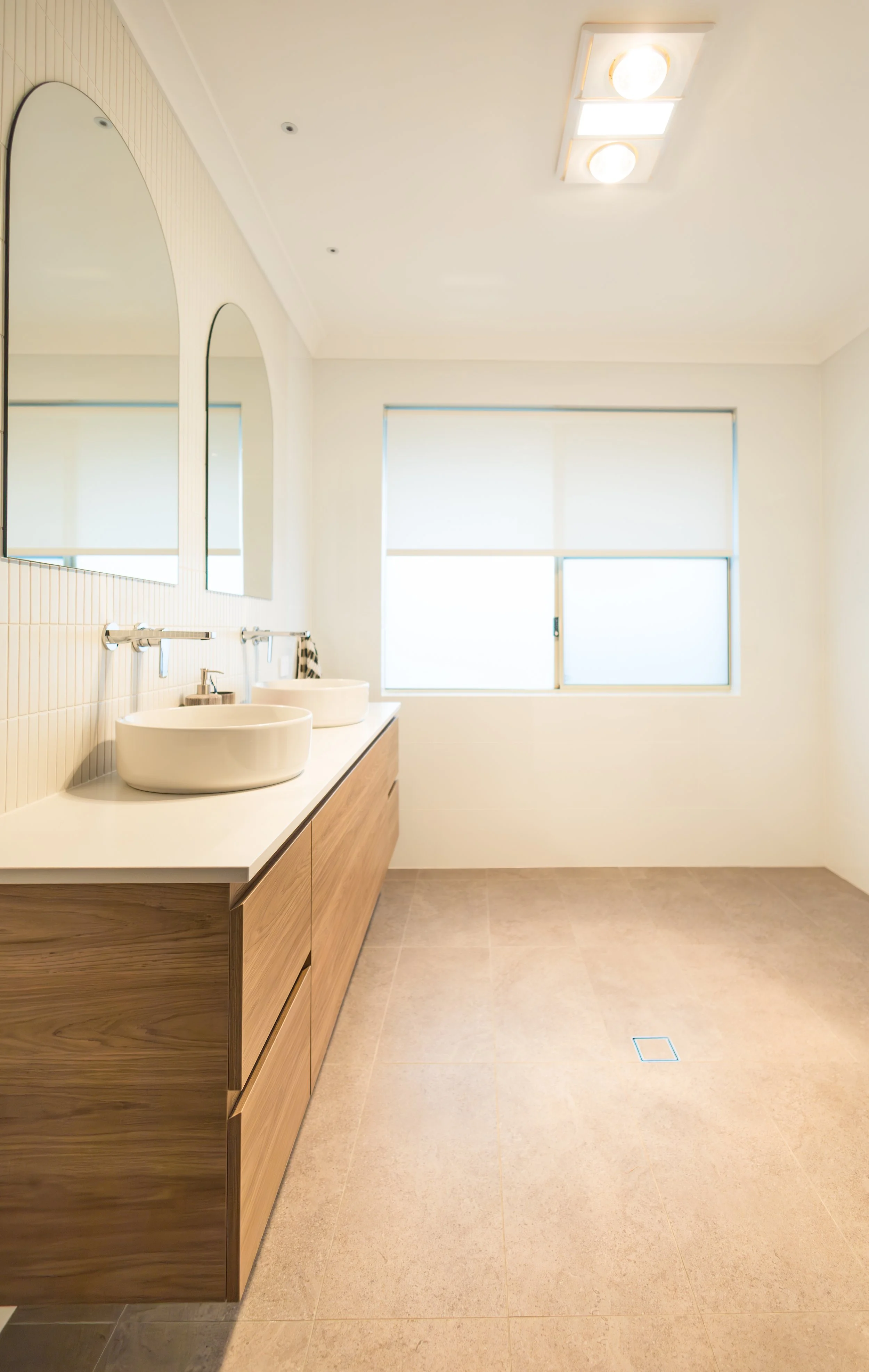 Modern bathroom with two vessel sinks on a white countertop, large mirrors, a window with blinds, and beige tiled flooring.