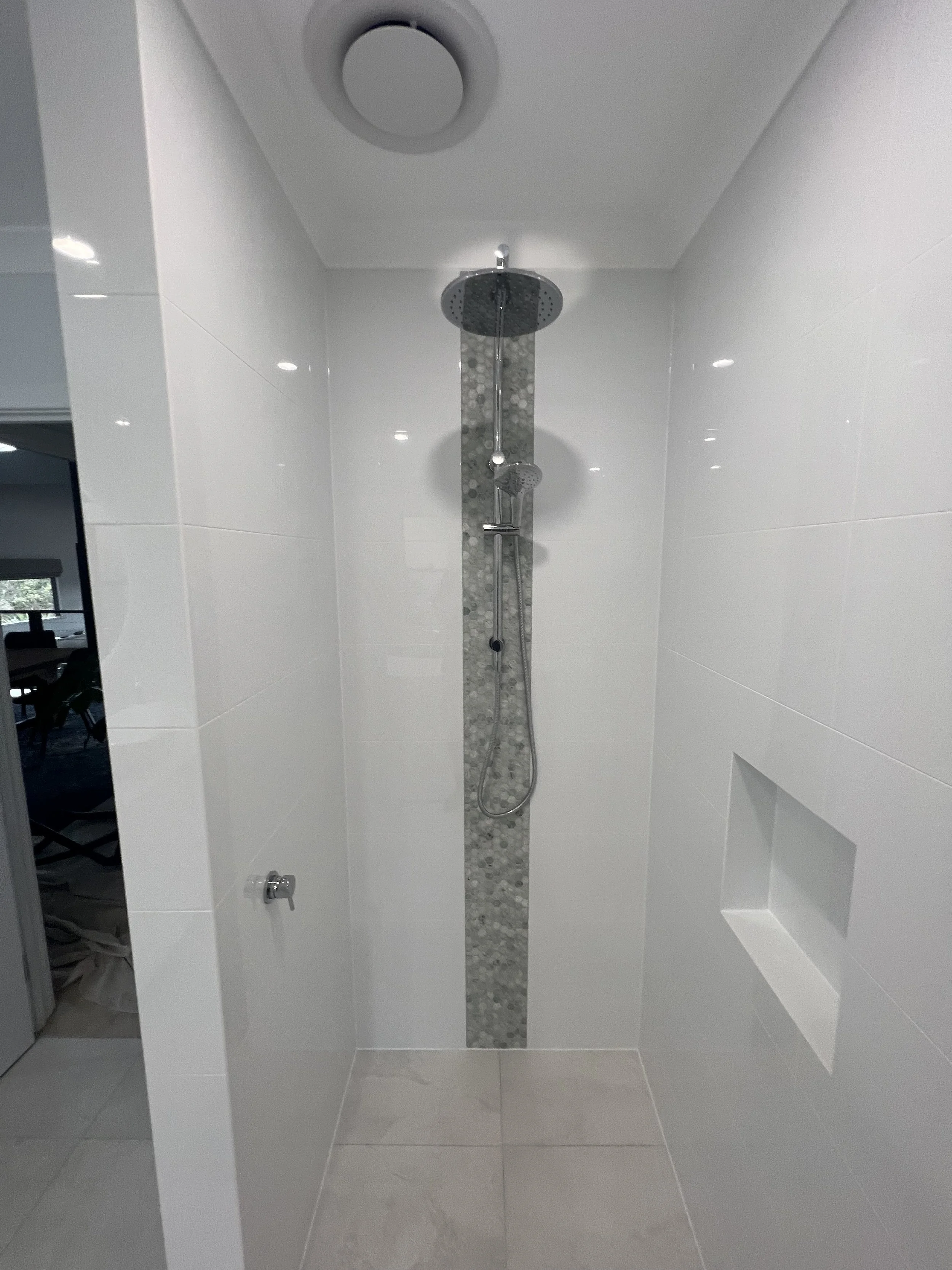 Modern walk-in shower with a rain shower head mounted on the ceiling and a handheld shower attached to a vertical strip of pebble-like tiles on the back wall, with a built-in niche on the right wall.