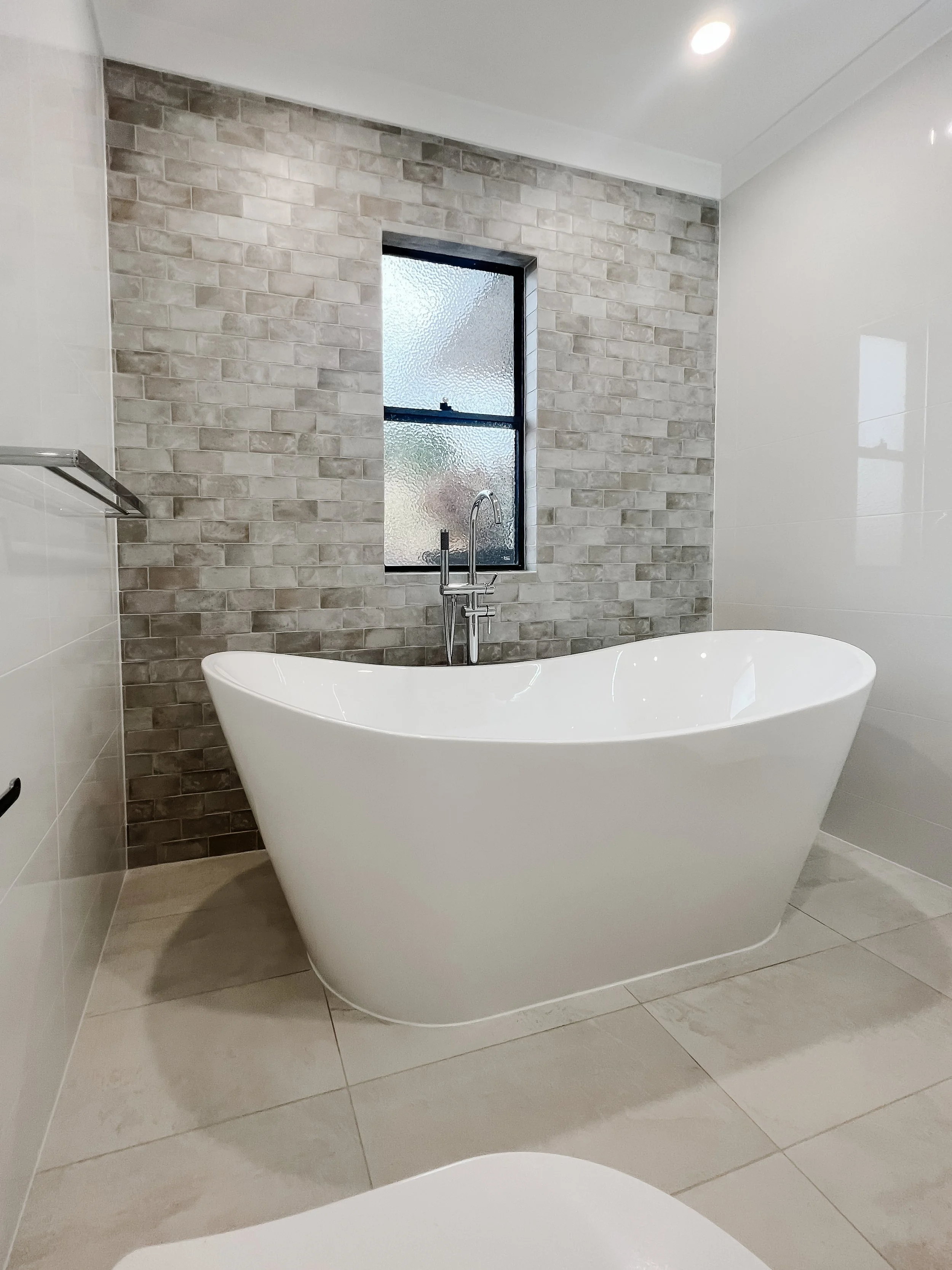Modern bathroom with a white freestanding bathtub against a brick accent wall, a window with frosted glass, and a ceiling light.