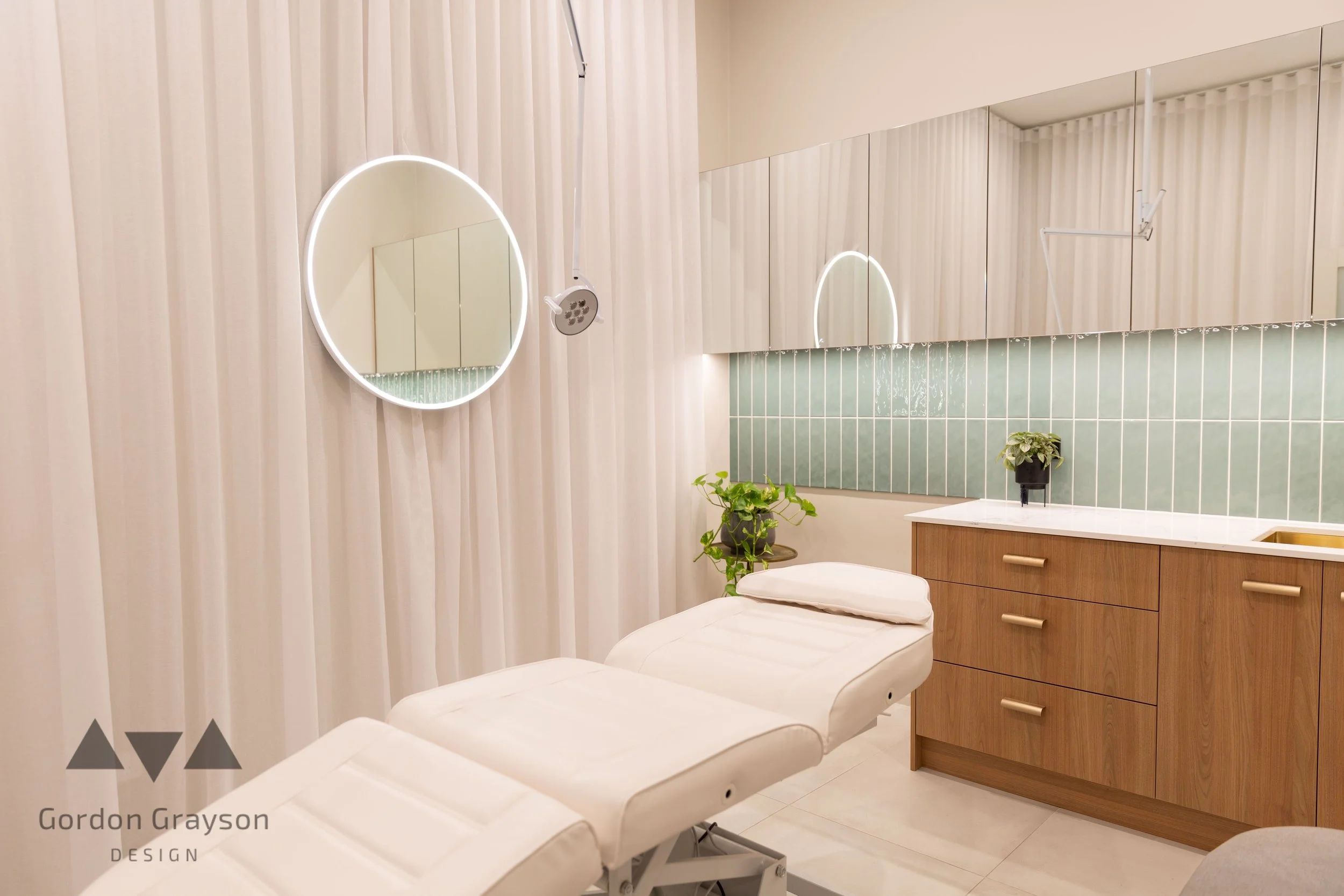 A clean, modern medical or spa treatment room with an adjustable white examination chair, a large mirror on the wall, green tiled backsplash, wooden cabinets by local custom builder in busselton