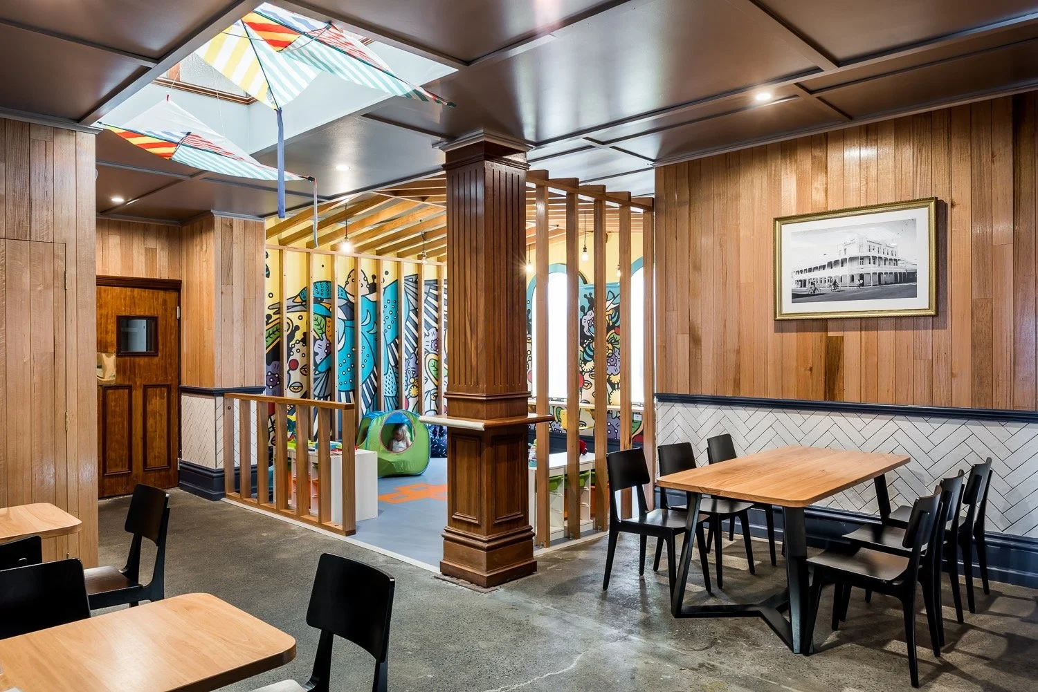 Indoor dining area with wooden walls, black chairs, and tables. A children’s play area with a colorful mural and toys is visible through wooden slats, with a skylight above. Commercial shop renovation and fit outs in western australia