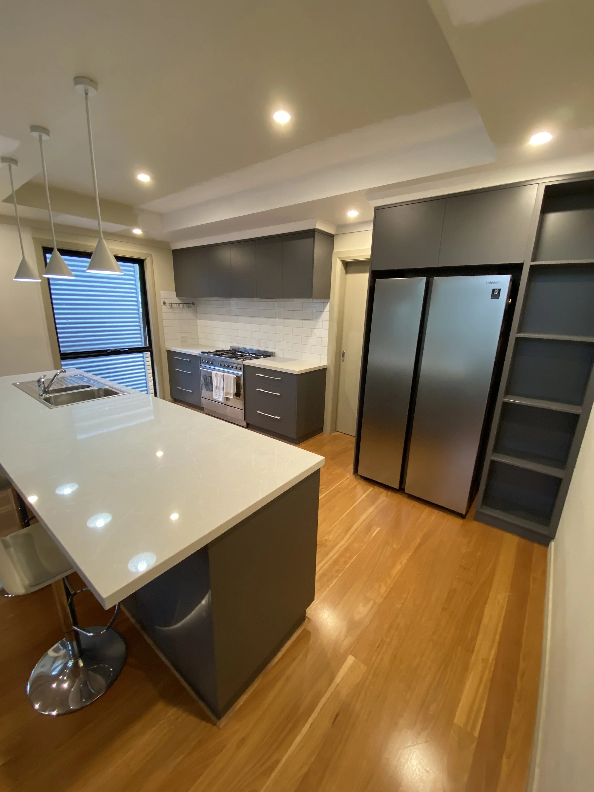 Modern kitchen with grey cabinets, white countertops, built-in oven, stainless steel refrigerator, white subway tile backsplash, wood flooring, and pendant lights overhead.