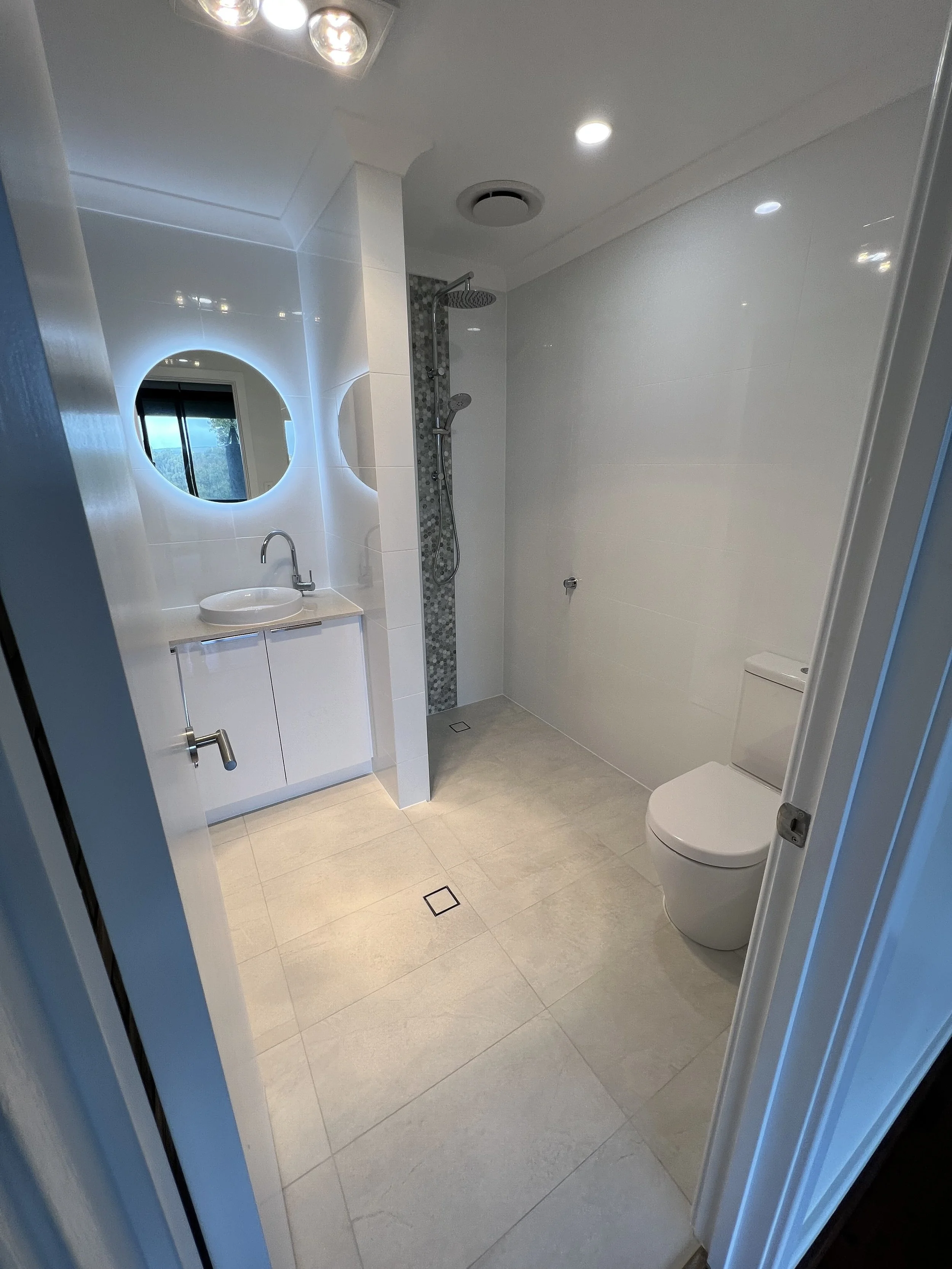 Modern bathroom with white walls, light-colored tile flooring, a floating vanity with an oval sink, a circular backlit mirror, a shower area with a rain showerhead and a handheld shower, as part of new renovation by local builder