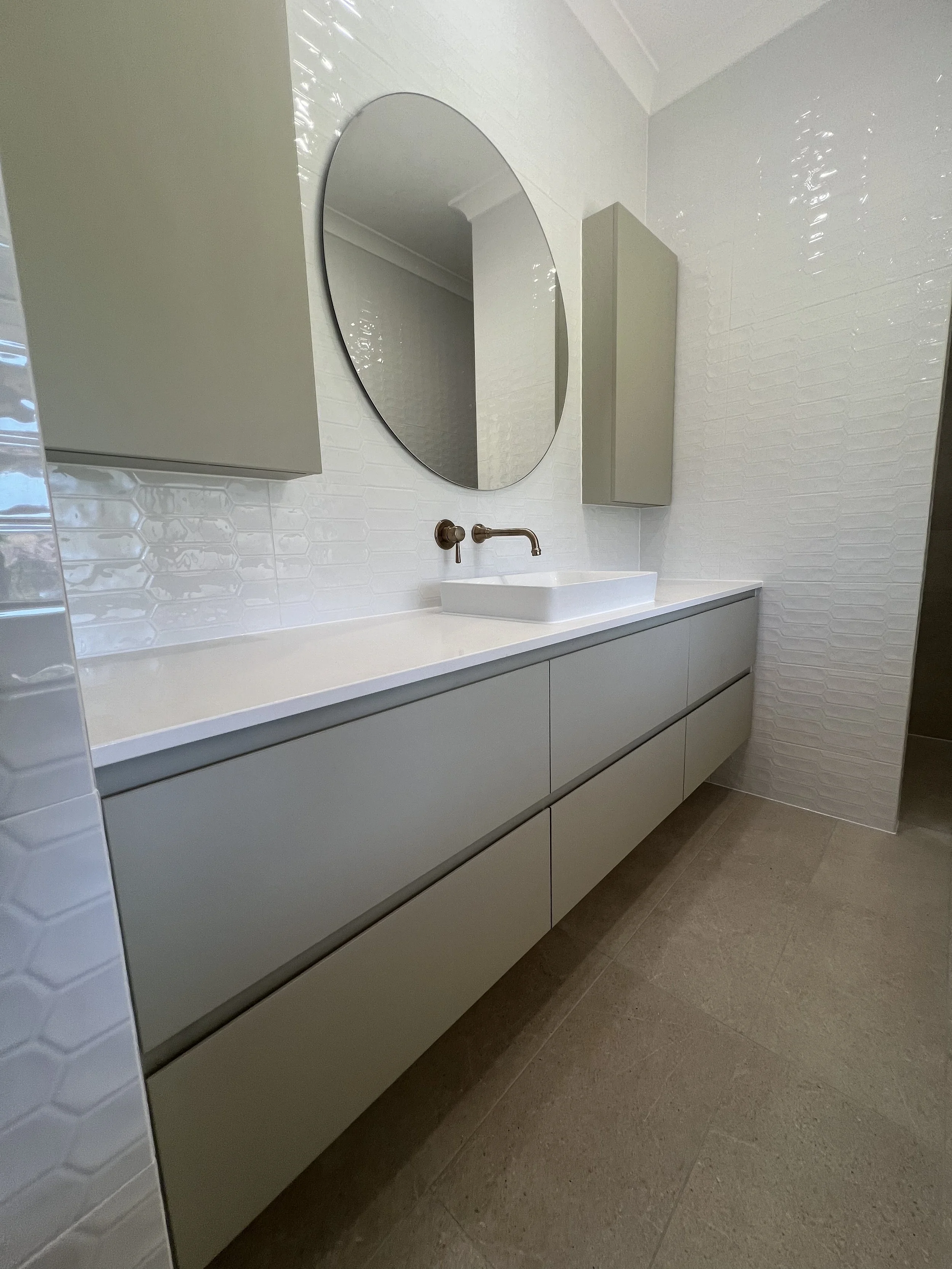 Modern bathroom vanity with light gray cabinets, a white countertop, a rectangular vessel sink, a wall-mounted faucet, an oval mirror, and wall cabinets on white textured tile walls.