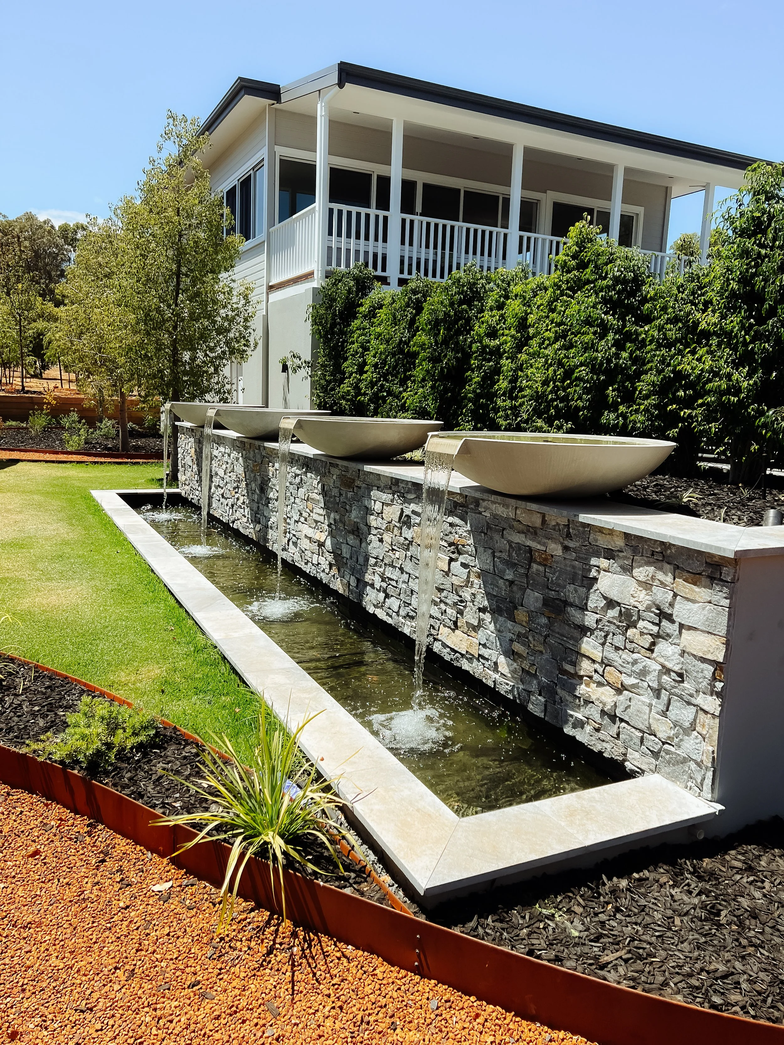 A backyard water feature with three bowls pouring water into a rectangular pool, located in front of a multi-story house with a balcony. The area is landscaped with green grass, shrubs, and mulch.