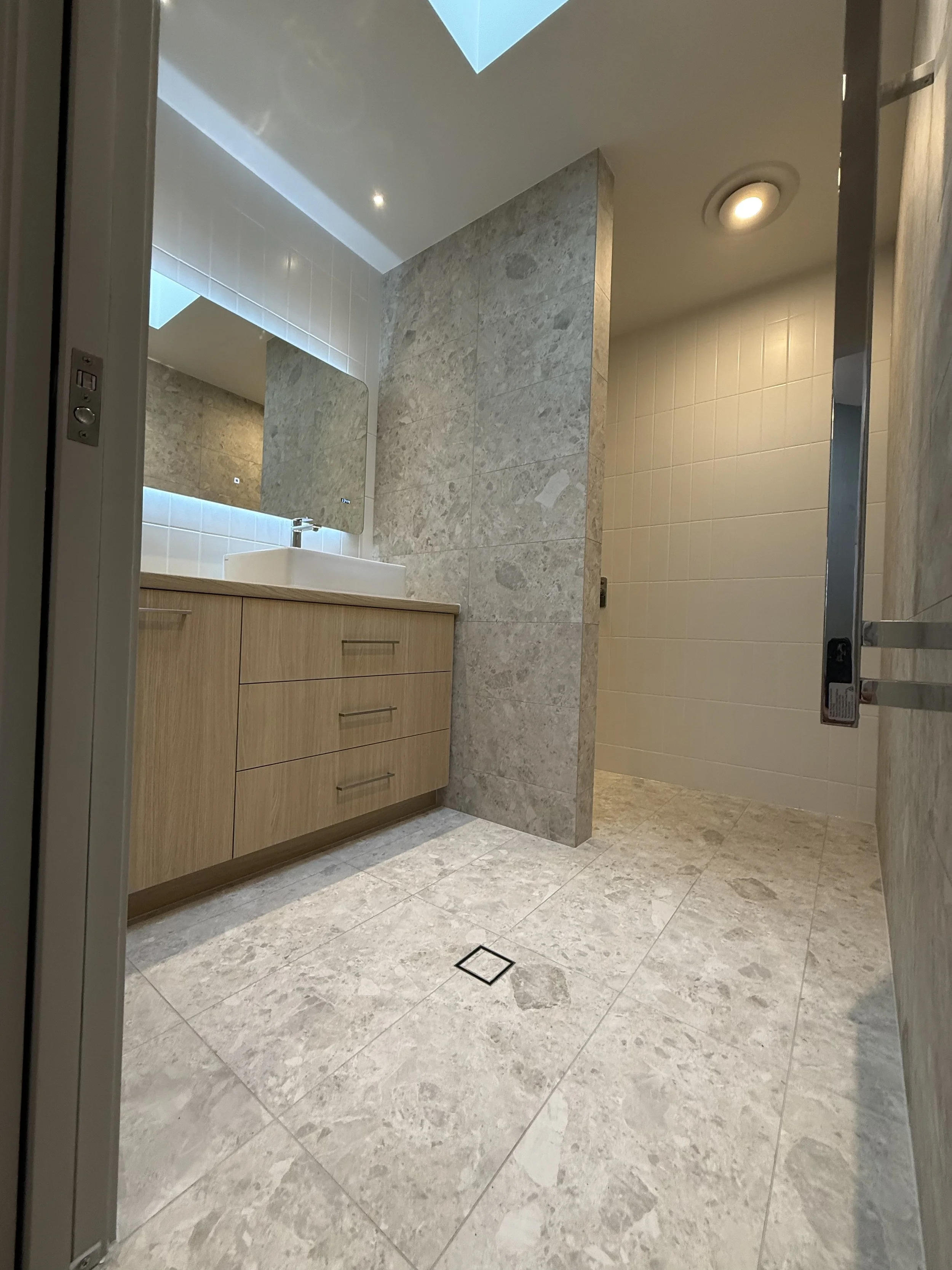 Modern bathroom with a wooden vanity, rectangular vessel sink, large mirror, tiled walls and floor, skylight, and recessed ceiling light.